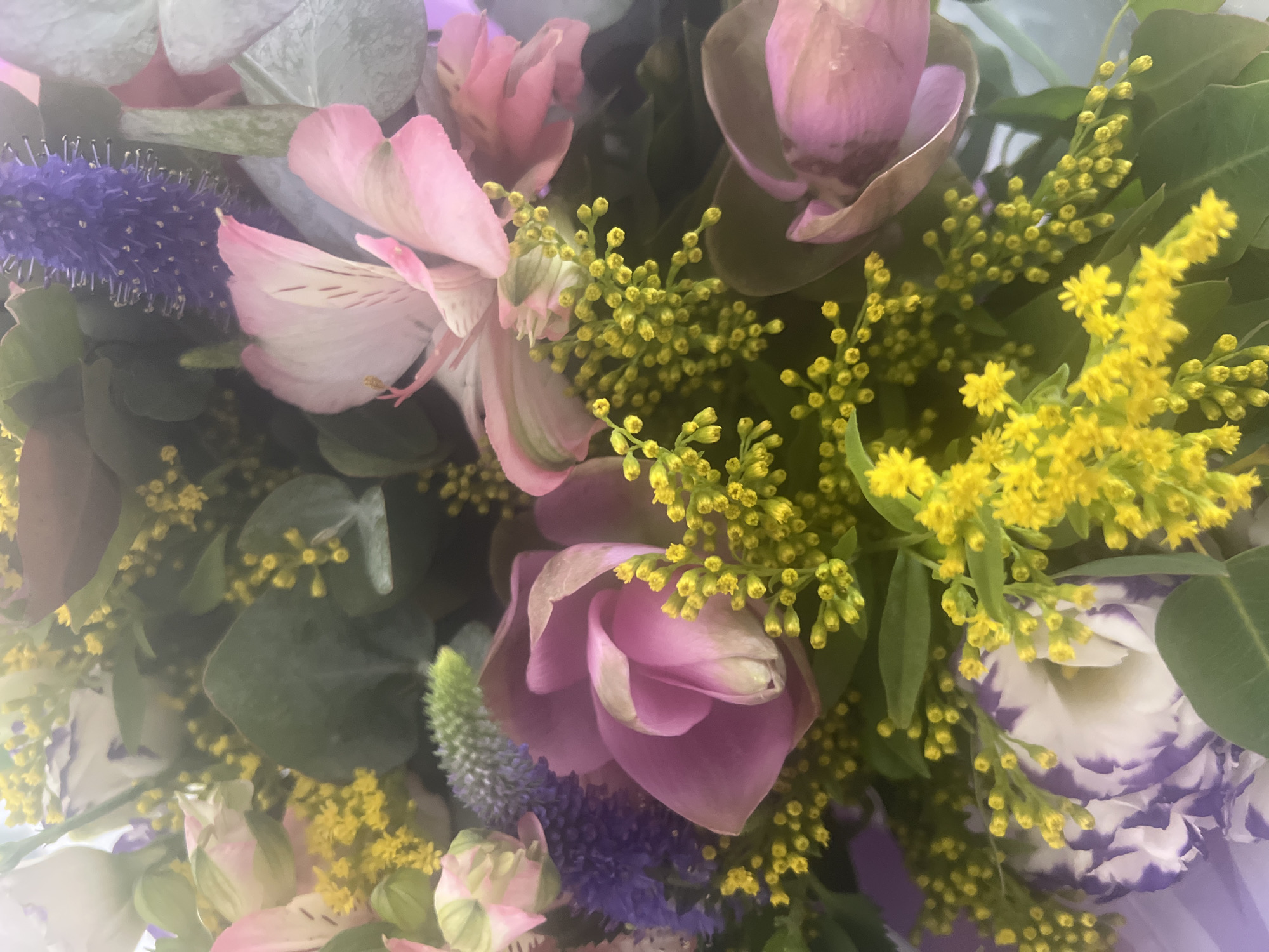 A close-up, overhead shot captures a vibrant bouquet of flowers, predominantly featuring soft pink alstroemeria, accented with delicate purple blossoms and interspersed with clusters of small yellow flowers and green foliage. The bouquet, likely arranged for an event or as a gift, is professionally composed. The image does not contain any identifiable landmarks, people, or actions, and thus cannot be definitively located within Hof HaCarmel Regional Council, Israel. The lighting appears to be natural or studio-based, creating a soft focus that emphasizes the textures and colors of the flowers.