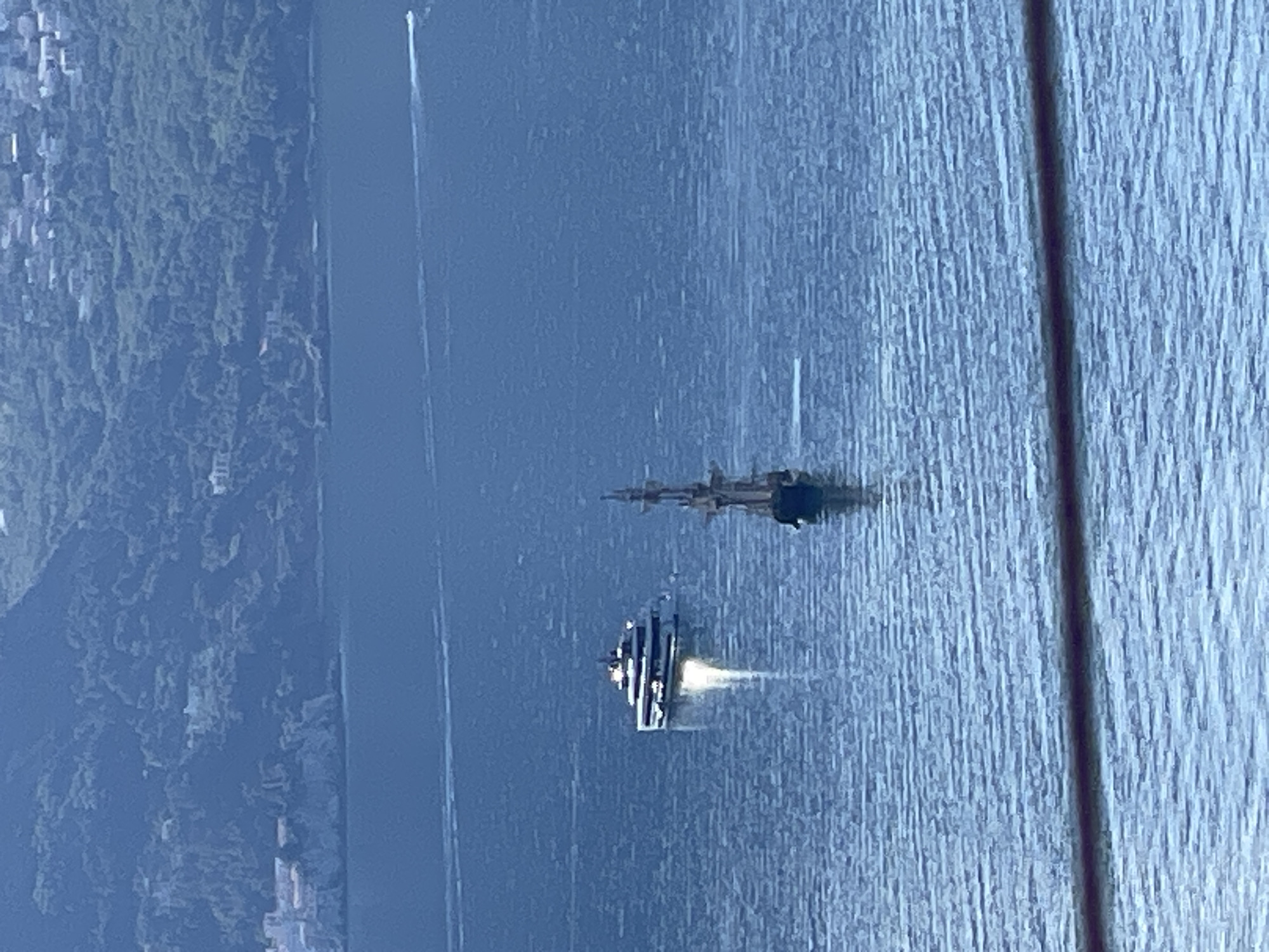 The image shows a view of the sea near Budva, Montenegro, from an elevated perspective. A large, modern yacht is visible, leaving a wake behind it. Further in the distance, another vessel, possibly a sailboat, is present. On the left of the image, the land with vegetation and buildings can be seen. A cable or wire runs vertically down the right side of the frame, likely a part of a structure from where the picture was taken.