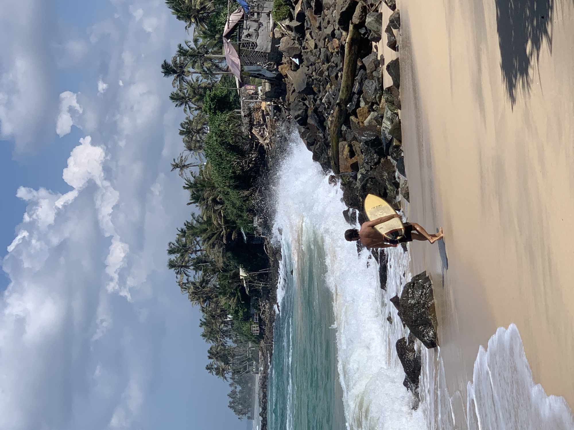A medium-shot photo of a tropical beach scene in Ahangama, Sri Lanka, depicts a tan-skinned person standing on the sandy shore holding a yellow surfboard. The individual is positioned near dark rocks, where waves are crashing, indicating the boundary between land and sea. The person appears to be preparing to enter the water.
Behind the person and the rocks, lush tropical vegetation and palm trees are visible, suggesting a vibrant coastal ecosystem. Further back, simple structures and buildings are nestled among the trees, possibly indicating a small village or settlement. The sky is a blend of blue and clouds, offering a dynamic backdrop to the coastal scene. The overall composition of the image captures a moment of anticipation and connection with the natural environment.