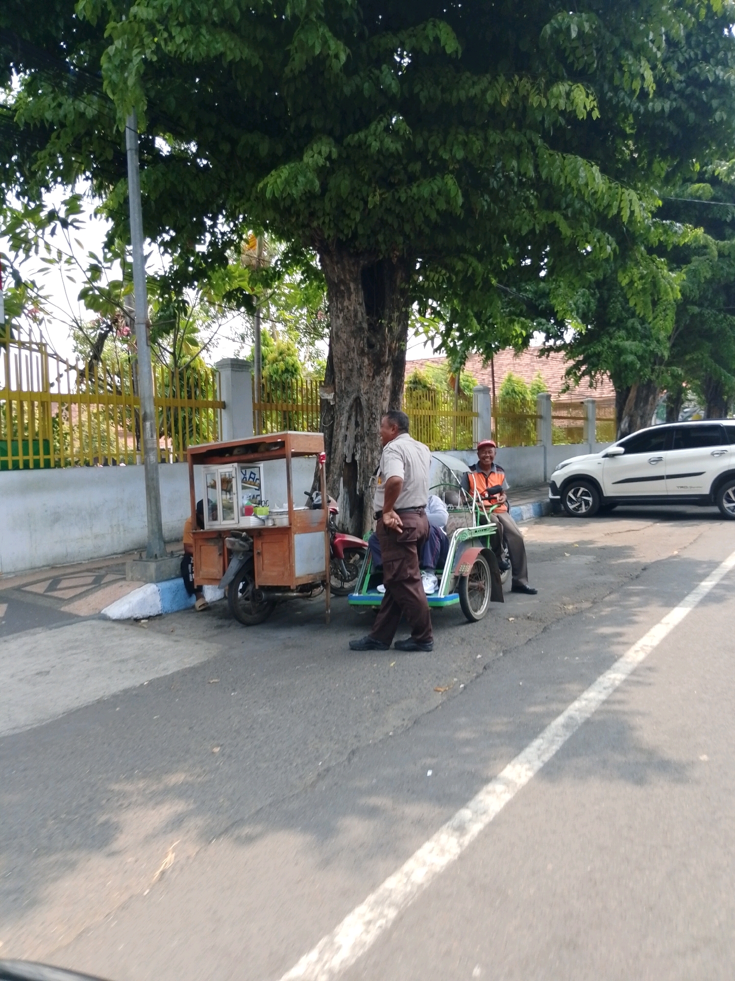 The image depicts a street scene in Pasuruan, Indonesia, with a focus on two men and a food cart. A mature, large tree with dense green foliage dominates the upper portion of the frame, casting shadows on the asphalt road. To the left, a yellow metal fence with vertical bars runs along a light-colored wall.
In the foreground, a three-wheeled food cart, appearing to be made of wood with metal accents, is parked on the side of the road. It has a display window and shelving, suggesting it is used for selling food or drinks. A red motorcycle is attached to the rear of the cart.
Two men are present. The man on the left, wearing a light-colored collared shirt and brown pants, stands with his hands behind his back, looking to his left. He appears to be an older gentleman. To the right, another man, wearing an orange vest over a light-colored shirt and brown pants, is seated in a small, green passenger carriage that is part of a modified bicycle or rickshaw-like vehicle. He is looking towards the other man and smiling. Another person, partially obscured, is visible in the passenger carriage with him.
In the background, a white SUV is parked on the right side of the road. The environment suggests a typical urban or semi-urban street with trees, buildings, and a road marked with white lines. The overall lighting indicates it is daytime.