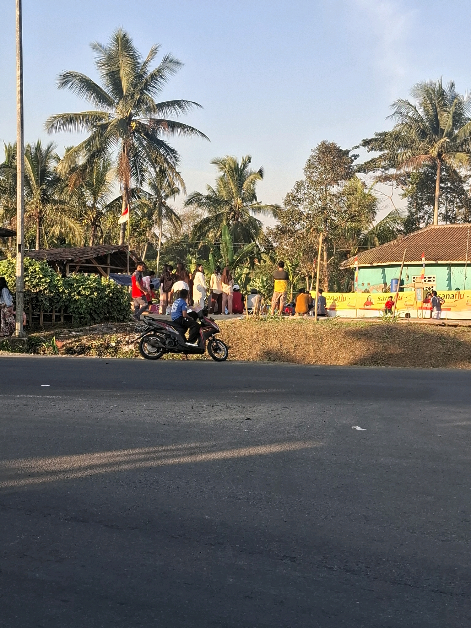 The image captures a street scene in Cikaret, Indonesia, under a clear sky. A group of people is gathered on the side of the road, some standing, some sitting. Among them, a person is riding a black scooter. In the background, a light green building with a brown roof is visible, along with palm trees and other vegetation. A yellow banner with red text is displayed near the building. The road in the foreground is asphalt. The lighting suggests it is either early morning or late afternoon, with long shadows cast across the road.