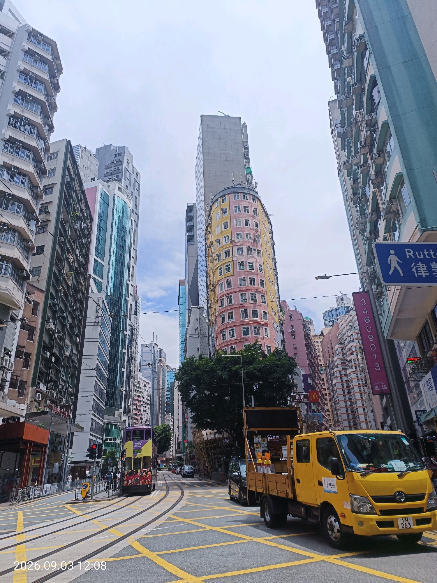 The image depicts a busy street scene in a dense urban environment, likely in Hong Kong, given the presence of trams and the architectural style. The perspective is low-angle, looking up at towering buildings on either side of a wide street. Yellow tram tracks are prominently visible in the foreground, with a yellow truck driving on the right side of the road. A purple and yellow double-decker tram is moving away from the viewer on the tram tracks. The buildings are a mix of modern skyscrapers and older, more compact residential blocks. Some buildings feature prominent air conditioning units on their exteriors. Signage in Chinese characters is visible on several buildings. The sky is overcast. The scene appears to be captured during the daytime. A timestamp in the bottom left corner reads "2026.09.03 12:08".