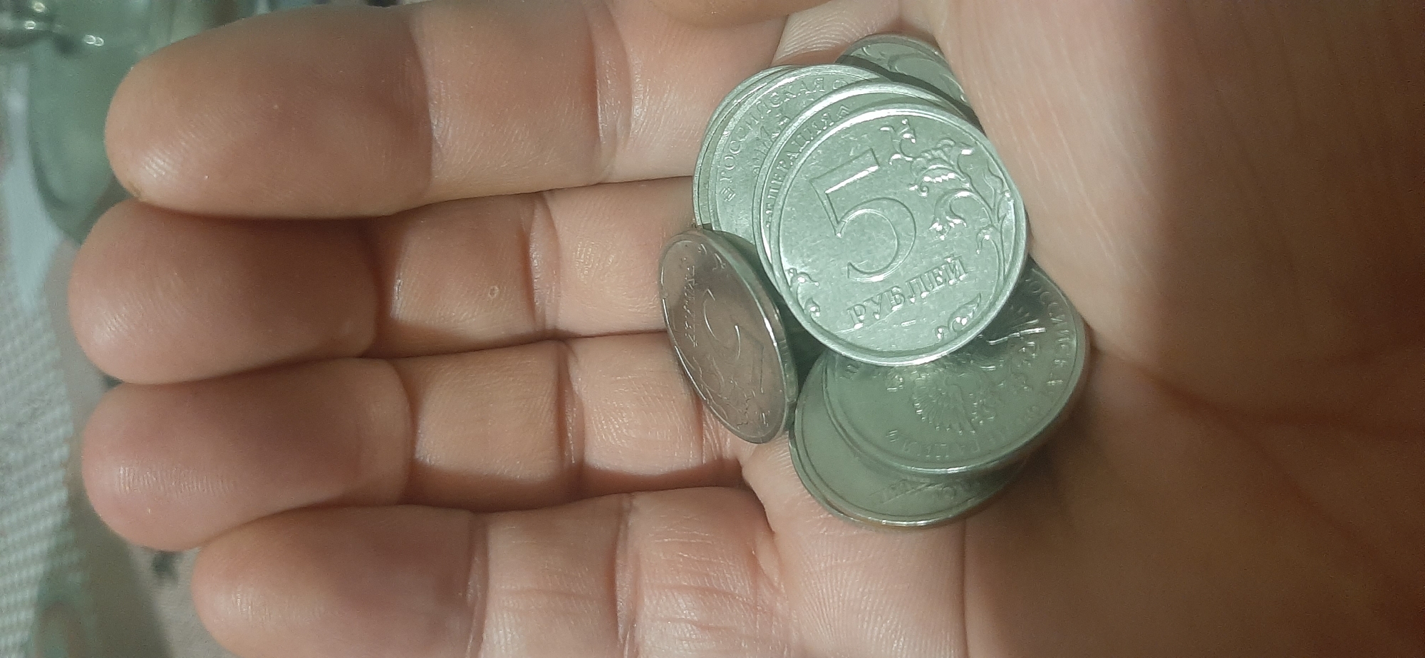 A close-up, vertical, overhead shot shows a person's hand holding several five-ruble coins in Yessentukskaya, Russia. The coins are a silvery-gray color and are stacked in a pile. The hand is open, with the fingers curved slightly to cradle the coins. The background is a blurry, patterned surface. The lighting is dim.