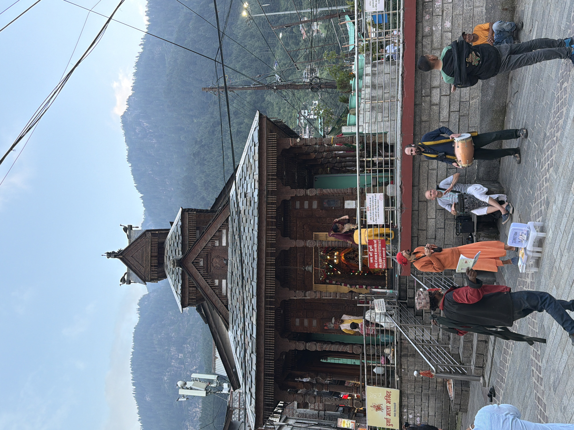 The image depicts a street scene in Manali, India, in front of a wooden temple with intricate carvings. A group of people are gathered, some of whom appear to be performers. In the foreground, a man in an orange robe with a red head covering stands with a book, possibly a performer or religious figure. To his left, another man in a checkered cap and red and black jacket is walking away, carrying a dark cloth object. To the right, two men are playing musical instruments: one is seated and playing a drum, while the other stands and plays a dholak. A young man in a black jacket and jeans is standing facing away from the camera. In the background, several people are seen near the entrance of the temple, including individuals in traditional attire. The temple itself has a stone base and a multi-tiered wooden roof. The backdrop features lush green mountains, characteristic of the Manali region. Power lines crisscross the sky, and streetlights are visible in the distance. The scene appears to be set during the late afternoon or early evening, with the sky overcast and a soft light illuminating the area. A sign with Hindi text is visible on the left side of the temple.