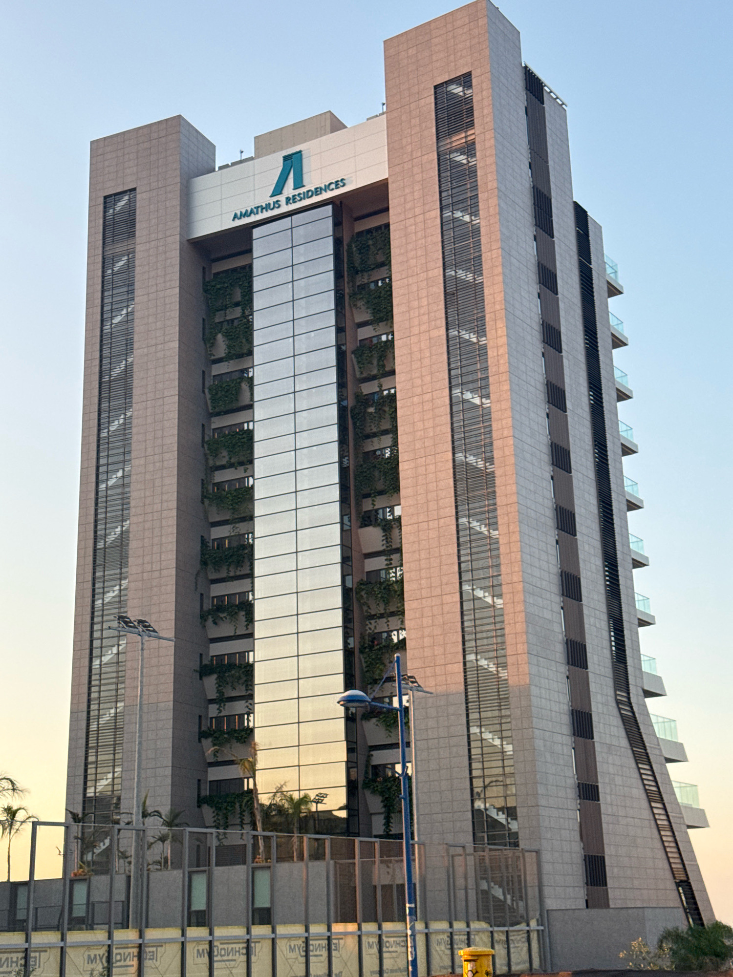 The image depicts the Amathus Residences, a modern, multi-story building located in the city of Agios Tychona Community, Cyprus. The structure features a prominent facade with a combination of beige stone cladding and large, reflective glass panels. Greenery, in the form of vertical gardens, is integrated into the design, cascading down the sides of the building. Balconies with glass railings are visible on the right side of the building. The sky above is clear and blue, with a hint of sunset glow on the horizon. In the foreground, there are streetlights and some palm trees, suggesting an outdoor urban environment. The bottom of the image shows a section of what appears to be construction fencing or temporary barriers, with some text on them.