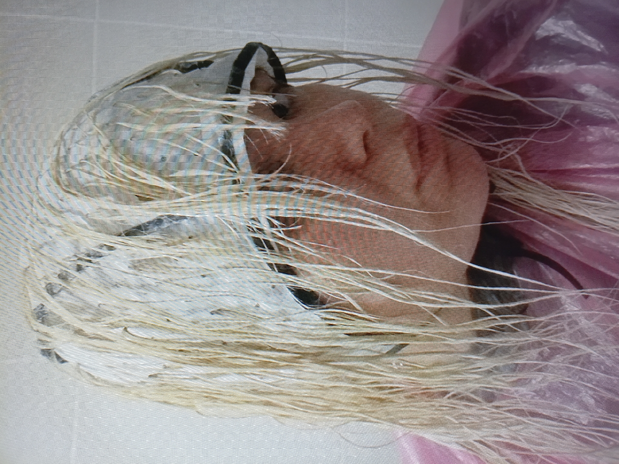 The image shows a close-up, head-and-shoulders shot of a person with blonde, wet hair. The hair is partially covered by a white cap with a patterned design. The person is wearing a pink, plastic cape. The background is a plain, light-colored wall, suggesting an indoor setting like a bathroom or salon. The lighting is flat, and the image quality is poor, with visible pixelation. There is no indication of the city of Tiraspol, Moldova, or any other specific location.
