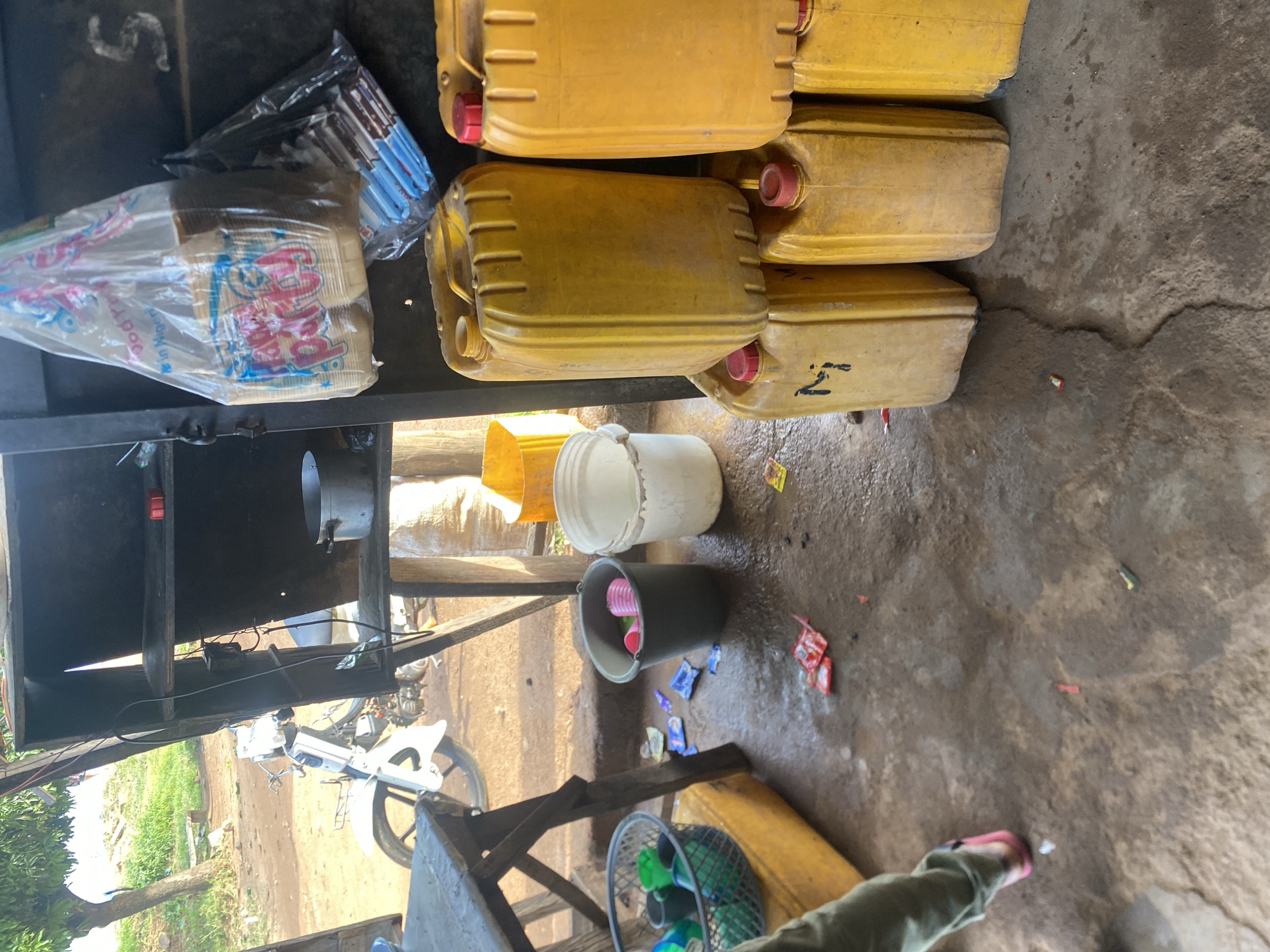 In Jos, Nigeria, a scene unfolds with a focus on a roadside stall or makeshift shop. The ground is a mix of dirt and wet patches, suggesting recent rain or water spillage. Scattered on the ground are small, colorful packets, possibly food wrappers or sachets. To the left, a person's lower legs and feet are visible, wearing a green garment and pink sandals, indicating their presence within the frame.
Dominating the right side of the image are several large, yellow plastic jerrycans, stacked in a way that suggests they are for sale or storage, likely of liquids. Some have red caps, and one has "Nj" scrawled on it. In the background, a white motorcycle is parked, hinting at a transportation hub or a nearby residence. Above the jerrycans, a plastic bag hangs, containing what appear to be paper cups or plates, some with colorful designs and text. Next to it, another dark plastic bag holds a bundle of blue and white items, potentially pens or straws.
Behind these items, shelves are visible, sparsely stocked with a silver pot on one shelf, and a small red object on another. The overall environment appears to be outdoors, with vegetation and trees visible in the distance, under a bright, possibly overcast, sky. The stall itself seems to be constructed from wood and metal, providing basic shelter.