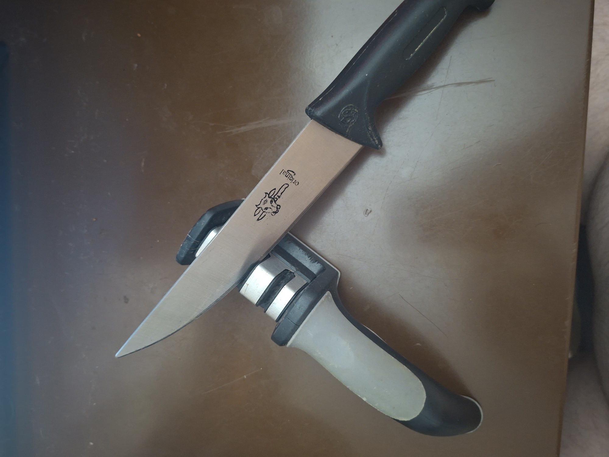 A metal knife with a black handle and a silver blade is positioned at an angle, with its tip resting on a knife sharpener. The knife sharpener has a grey and black handle and two silver sharpening grooves. The objects are on a brown surface, possibly a table. There is a faint image of a cow and the word "original" printed on the blade of the knife. The lighting in the scene is direct, casting shadows. This image was taken indoors.
