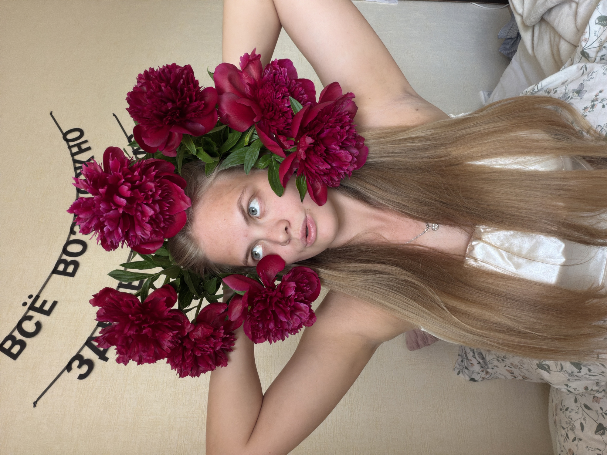 A young woman is photographed in a medium shot, lying down and looking upwards towards the camera. She wears a vibrant crown of numerous deep red peonies with green leaves encircling her head. Her long, straight light brown hair flows around her, spreading across the surface she is on. Her eyes are wide and light blue, and her mouth is slightly agape, suggesting an expression of surprise or playful wonder. She is wearing a light-colored top and a delicate silver necklace with a small, circular pendant. Her right arm is bent and raised above her head.
The background features a light beige wall on the left, displaying stylized black text in Russian, "ВСЁ ВОЗМОЖНО," which translates to "EVERYTHING IS POSSIBLE." On the right, a bed is visible with light-colored bedding, including a floral-patterned duvet or blanket and a plain white sheet. The setting appears to be indoors, likely a bedroom, with soft, diffused lighting. The image was captured in Izhevsk, Russia.