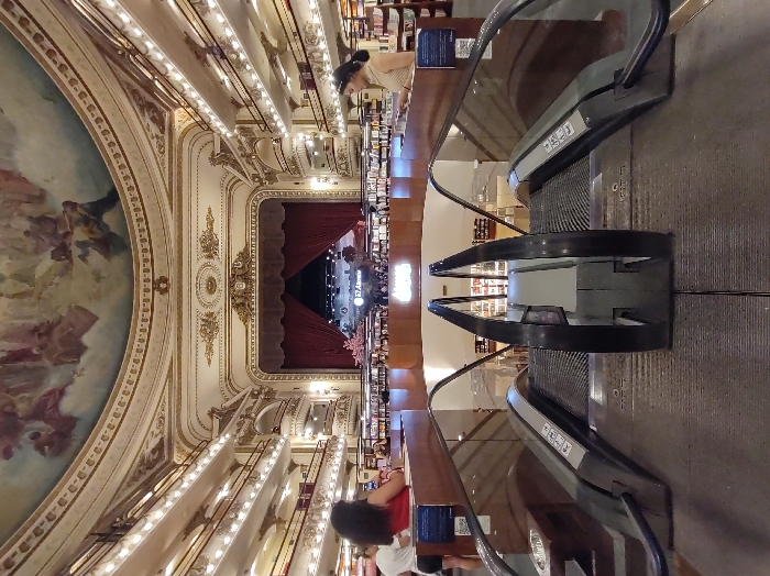 A view of El Ateneo Grand Splendid bookstore, converted from a theater. The ceiling features a large, ornate mural. The architecture includes gilded details and balcony boxes. Escalators run through the bookstore, passing displays of books. People are browsing on multiple levels. Red curtains still hang where the stage used to be.