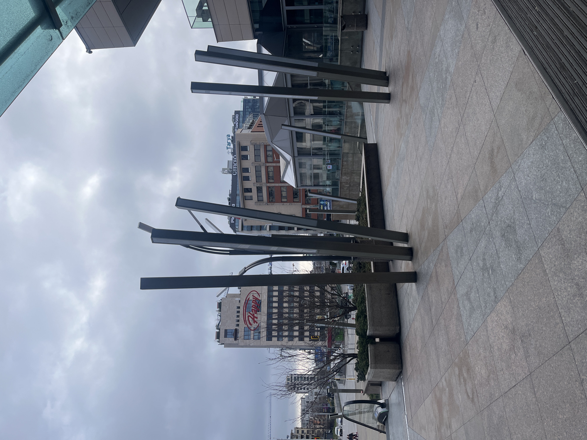 The image shows an outdoor urban scene in Sofia, Bulgaria. In the foreground is a modern art installation consisting of several dark metal vertical bars of varying heights and angles. The ground is paved with gray and tan tiles. Behind the installation, across the street, are several buildings. One is a tall, light-colored building with the word "HAPPY" in large red letters on its facade. Another building features the word "TARYA" on its top, while "OFFICE" is also visible on another part of the building. In the distance are more buildings and bare tree branches against a cloudy sky. Along the street, there are some concrete planters with greenery and hints of passing cars and pedestrians. The picture appears to be taken during the daytime under overcast conditions.