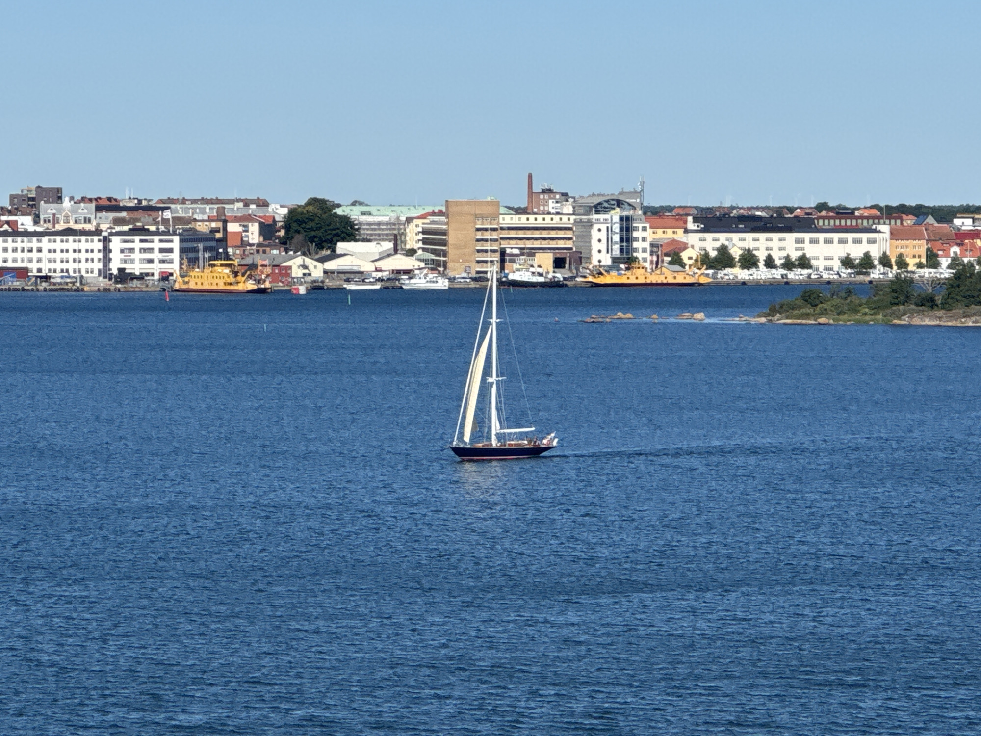 A wide shot captures a clear, blue sky above a cityscape on the coast of Karlskrona, Sweden. The city features a collection of buildings with varied architecture, including residential blocks, commercial structures, and industrial elements. A prominent yellow ferry is docked near a building with large windows. In the foreground, a sailboat with white sails navigates the blue water. The scene is set on a sunny day, with calm water reflecting the sky.