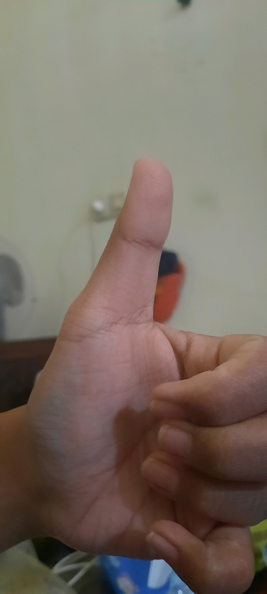The image shows a close-up of a person's right hand giving a thumbs-up gesture. The hand is in the foreground, with the thumb extended upwards and the other fingers partially curled. The skin tone is light. The background is blurred and appears to be an indoor setting with a light-colored wall. A partially visible red and black object is in the background, possibly a bin or a bag. The overall lighting is dim. The image was likely taken in Sidoarjo, Indonesia, but this cannot be definitively confirmed from the visual information alone.