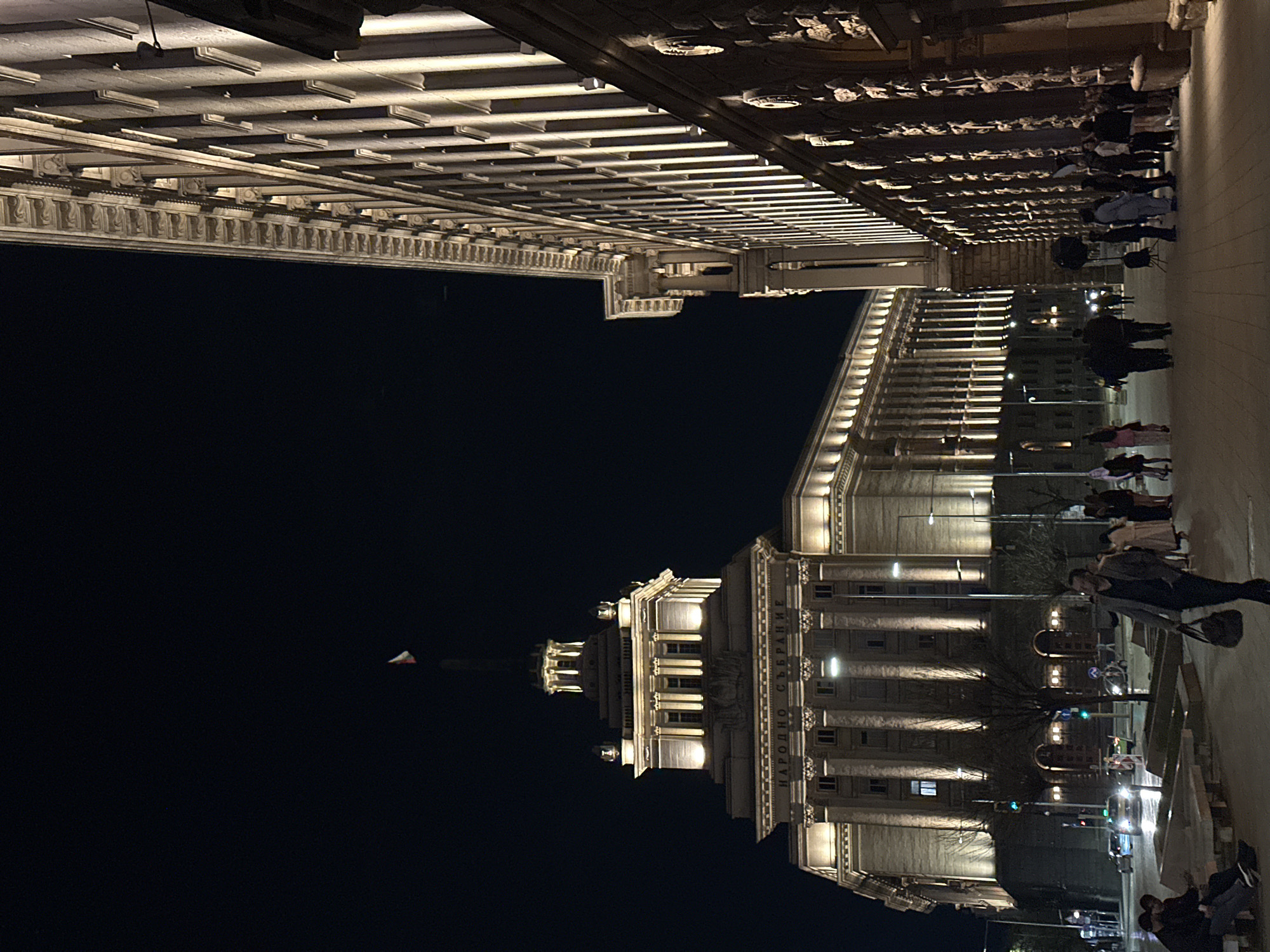 The image shows the city of Sofia, Bulgaria at night. The National Assembly building is illuminated on the right side of the image. On the upper portion of the image, there are architectural details of a building with lines of lights and the shadows of people along the walkway. The rest of the frame is dark, with some lights visible in the distance.