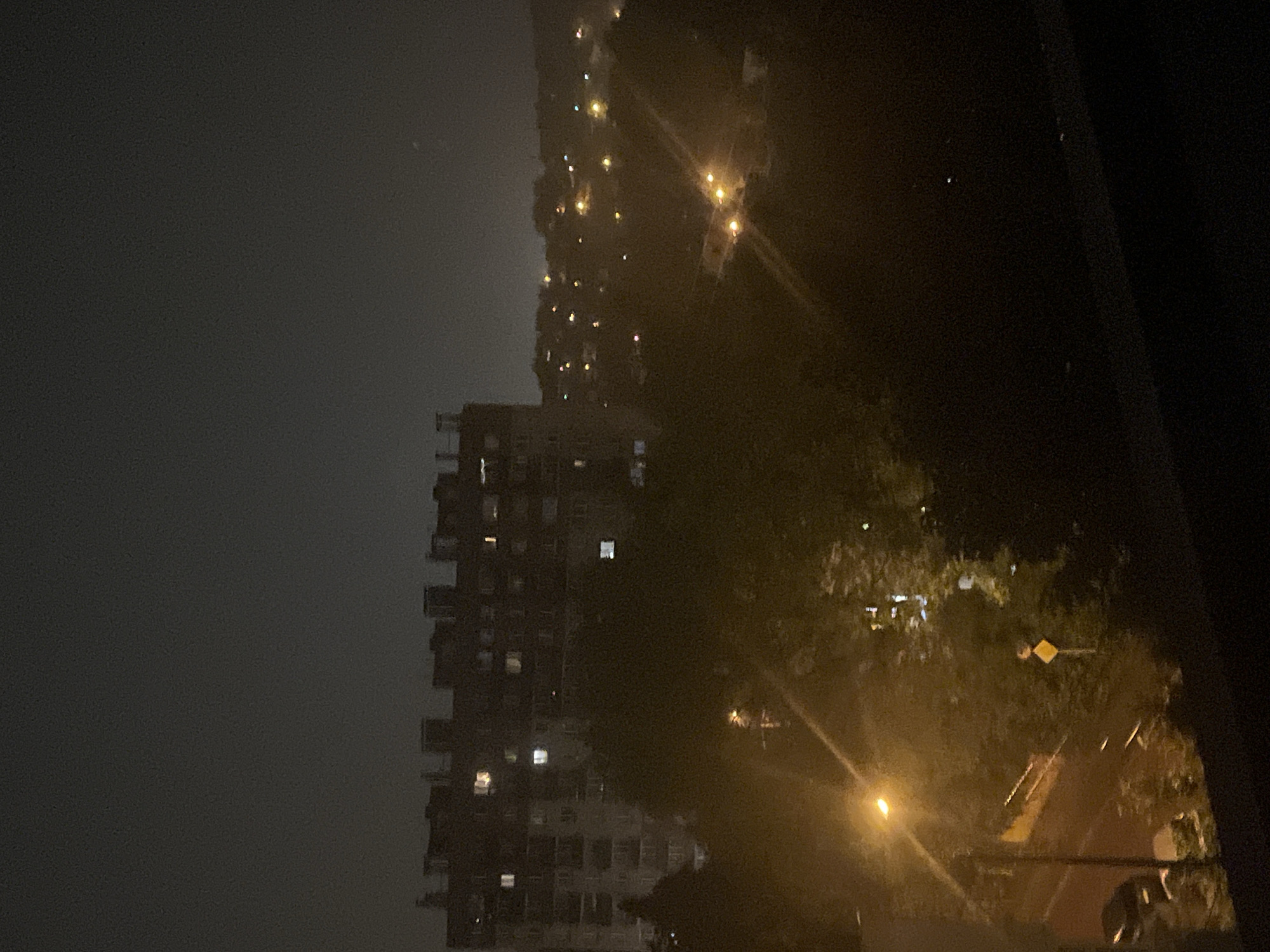 A nighttime aerial view captures a dense urban landscape under a dark, hazy sky. In the foreground, a dark railing or ledge runs across the bottom of the frame. To the left, a tall, multi-story apartment building is illuminated by scattered interior lights. Behind it, a dense cluster of trees obscures much of the midground. Further into the distance, a sprawling residential area is dotted with numerous small, glowing lights, indicating houses and streetlights. A prominent streetlamp with a bright orange glow illuminates a portion of a road and sidewalk in the lower left quadrant. A yellow diamond-shaped traffic sign is visible amidst the trees. The overall scene conveys a quiet, dimly lit urban environment at night, with a sense of depth created by the layering of buildings and vegetation.
