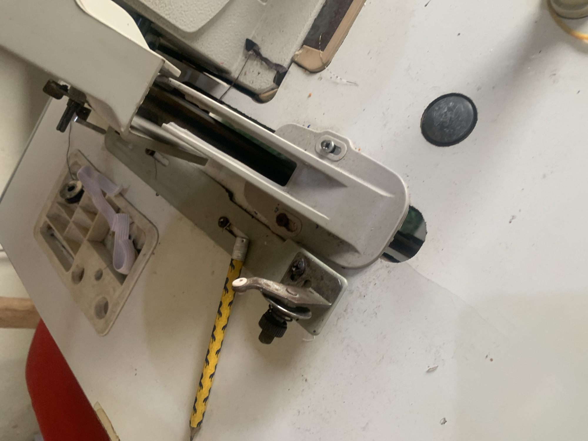 The image shows a close-up view of a sewing machine, specifically the area around the needle and presser foot. The sewing machine is white and appears to be well-used, with some dust and debris visible on its surface. A yellow and blue braided cord runs across the left side of the image, extending out of frame. A metal lever with a spring mechanism is attached to the machine's bed. The background is blurred, but appears to be an indoor setting with hints of red fabric and wooden elements. The scene does not contain any information that allows for localization to Jauro Yinu, Nigeria.