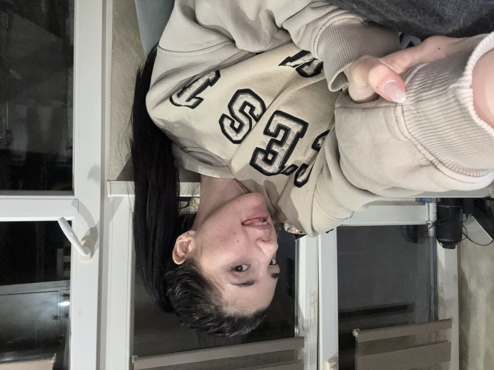 A young woman with dark hair, wearing a tan sweatshirt with the letters "ES" printed on it, is looking at the camera. She is inside, next to a window with a white handle. The room appears to be dimly lit. The location cannot be determined from the image alone. Sofia, Bulgaria cannot be determined from the image alone.