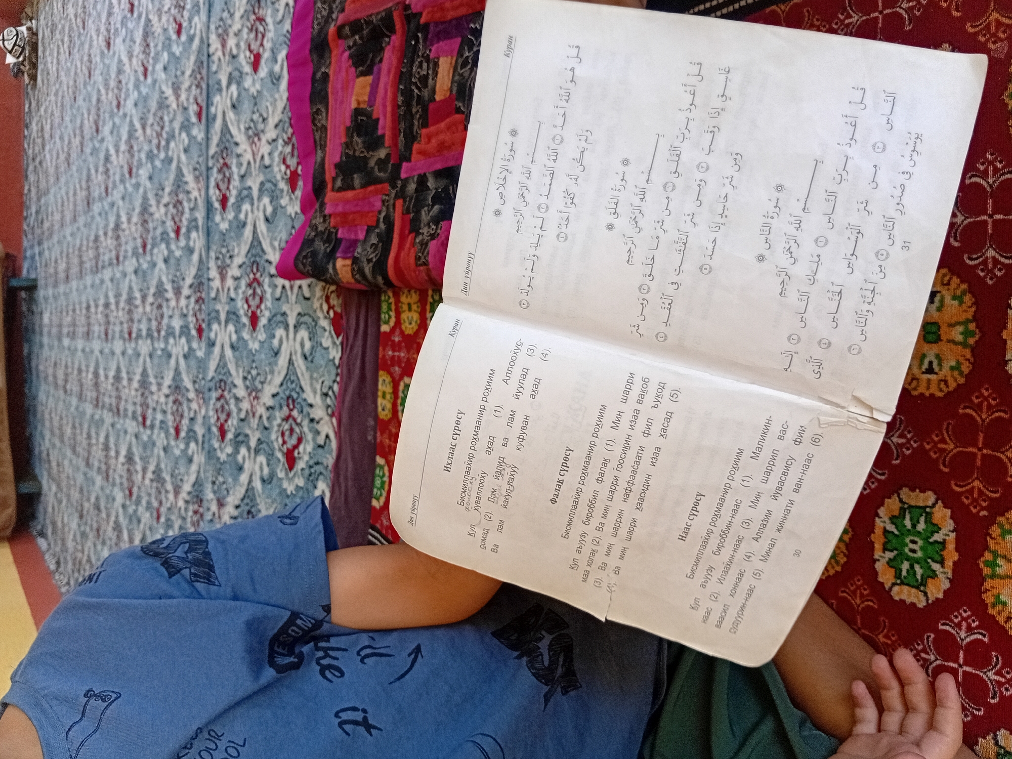 An overhead, close-up shot shows a child wearing a blue t-shirt with a graphic print. The child is sitting on a patterned rug in what appears to be an indoor setting, possibly a home. In front of the child, an open book is laid out on the rug. The book contains text in what seems to be a foreign language, with Arabic script visible on the right-hand page. The book also features illustrations and numbered verses. The rug itself has a complex geometric pattern in red, green, and white. To the right of the rug, a quilt with a patchwork of vibrant colors and patterns is visible. The lighting in the scene is natural, casting soft shadows. The overall impression is one of a candid moment, perhaps of a child engaged in reading or studying.