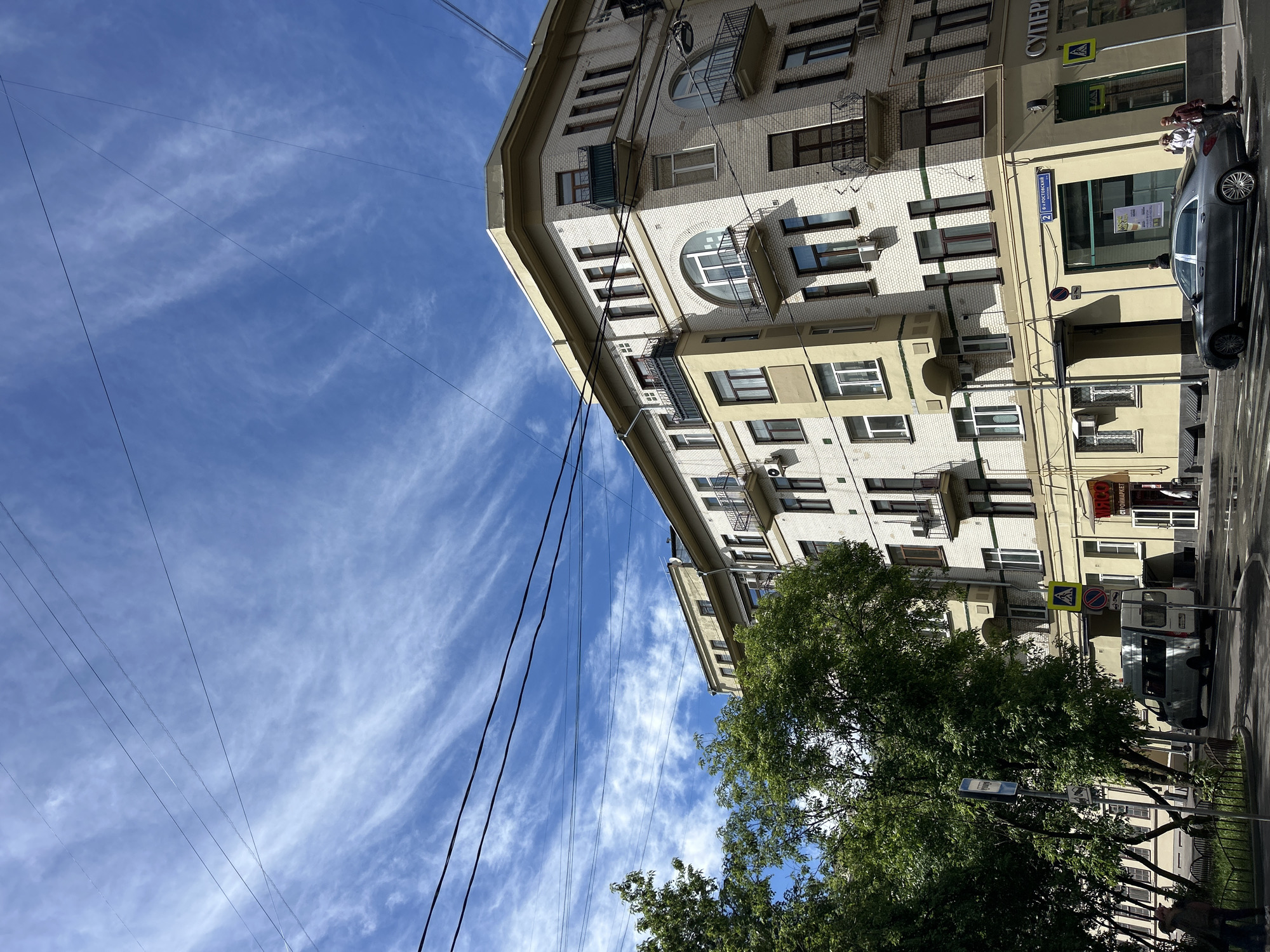 The image captures an urban scene in Unknown, Unknown, showing a building with light-colored brick facade, interspersed with cream-colored sections. The building features multiple windows, some with small balconies, and decorative archways. Street signs are visible near the building, including a pedestrian crossing sign and a street name plate "2 VOSTOCHNAYA".
A dark-colored vehicle is parked near the curb, with two individuals standing near it. Trees line the street, partially obscuring the lower portion of the building. Overhead, numerous power lines stretch across a blue sky marked with wispy clouds. The perspective of the shot is from a low angle, looking upward toward the building and sky.
