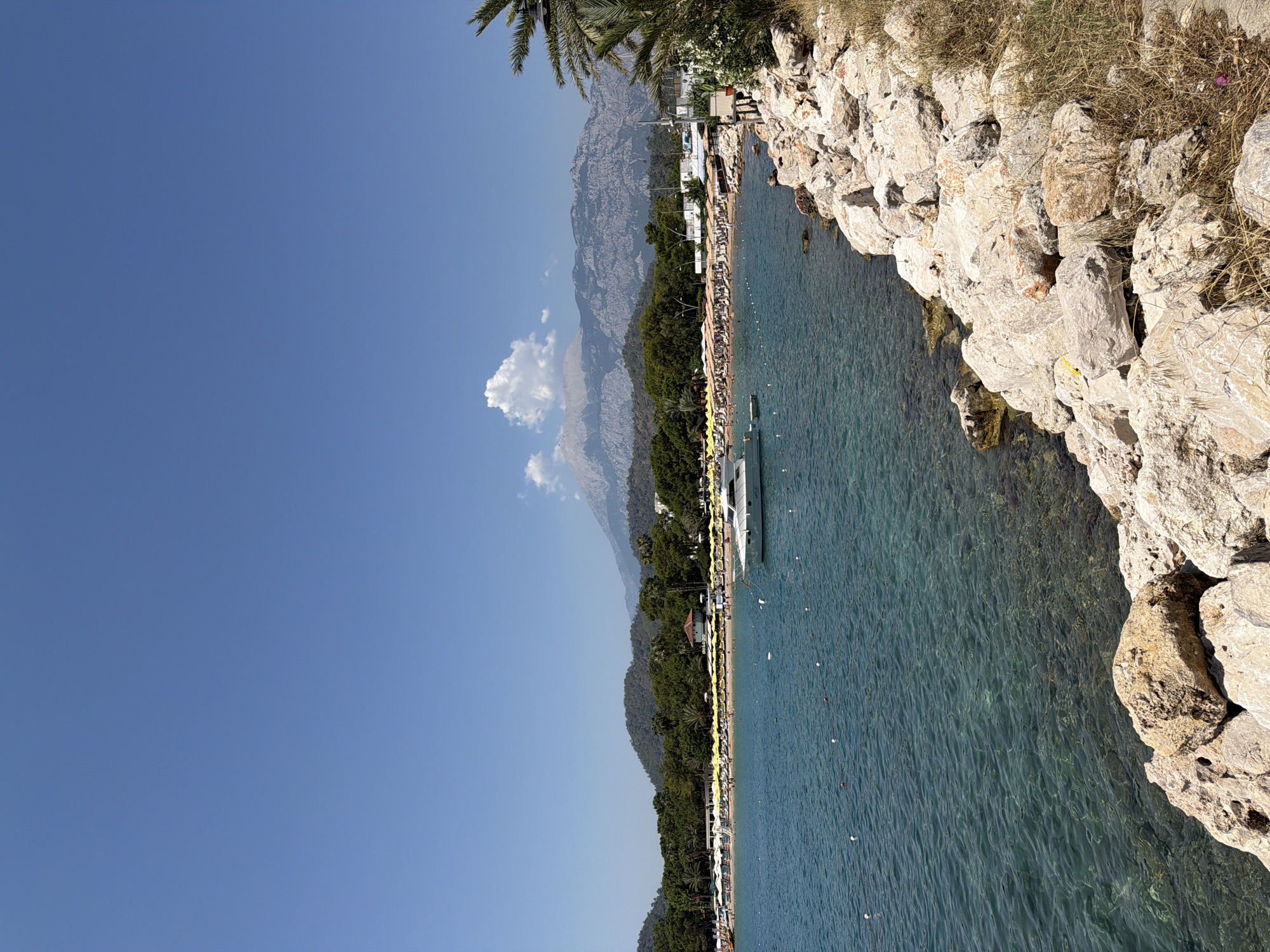 The image depicts a coastal scene with a clear blue sky. The foreground features a rocky shoreline with dry grass. The water is a vibrant turquoise, with a visible demarcation line of buoys stretching across it. In the middle ground, a white yacht is anchored offshore. Beyond the water lies a sandy beach lined with numerous yellow umbrellas and loungers, indicating a populated tourist area. A dense line of green trees forms a backdrop to the beach, with buildings visible interspersed amongst them. In the distance, a range of majestic mountains dominates the horizon, with a single large cloud formation present near one of the peaks. Palm trees are visible on the right side of the image, framing the scene. The overall impression is of a sunny day at a resort destination.