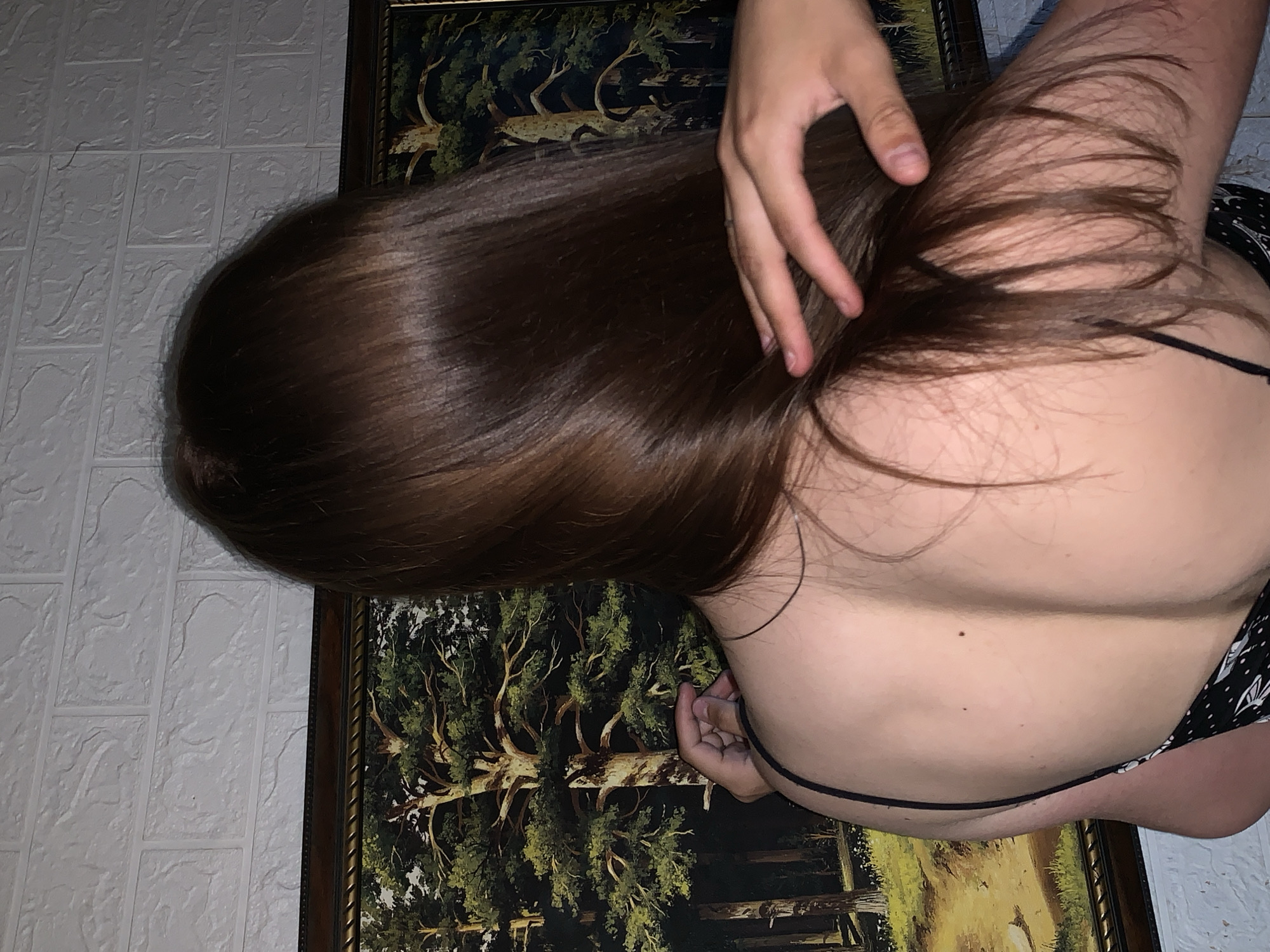 The image shows the back of a person with long, brown hair. They are wearing a black spaghetti strap dress with a white pattern. Their back is visible, with two moles on their upper back. A hand is reaching out from the right side of the frame, gently touching the person's hair. In the background, there is a framed painting of a forest with trees and a path. Above the painting is a white, textured wall that resembles brickwork. The scene appears to be indoors, possibly in a home or a room with decorative elements. The location in the city of Фоки, Russia is not discernible from the image itself.