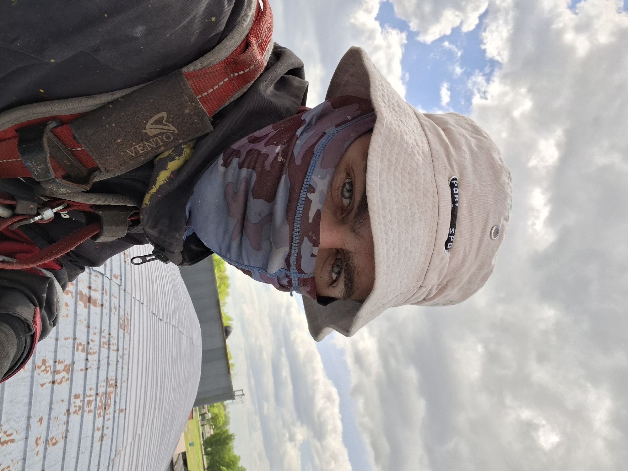 A person is wearing a safety harness with "VENTO" visible on a strap, a camouflage face covering, and a light-colored bucket hat with "PORT" visible on the side. The person appears to be standing on a metal roof. The background shows a cloudy sky, greenery, and what looks like a building or structure. The photograph was taken from a low angle, looking upwards. No verifiable location information is present within the image to confirm its location in the city of Zhabinka, Belarus.