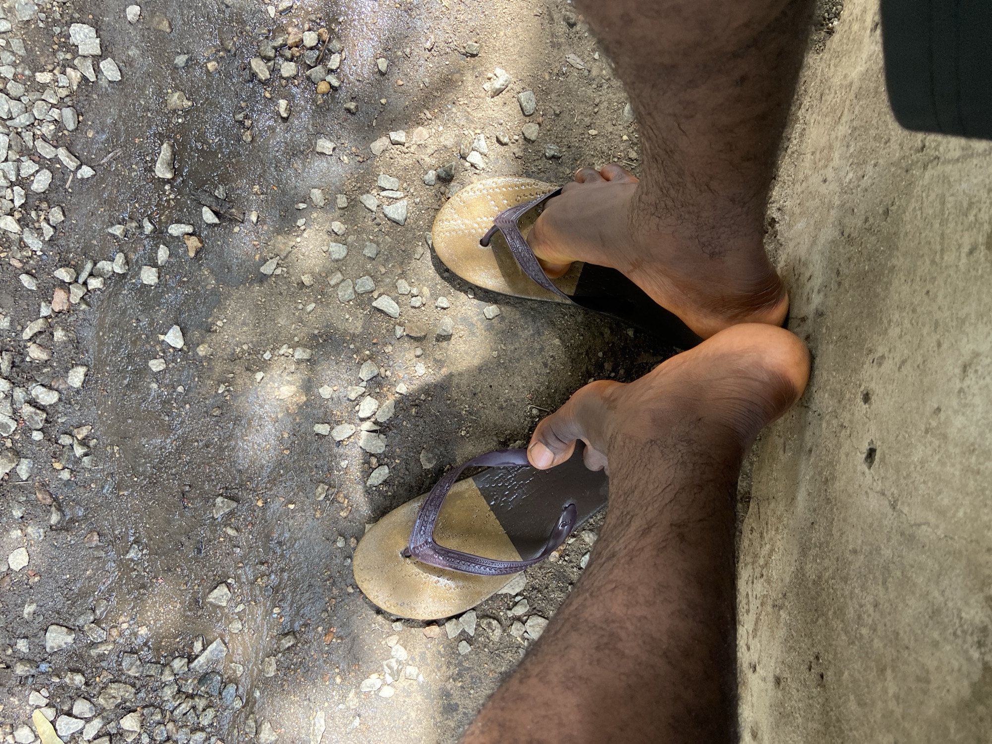 A first-person perspective photograph shows a pair of human feet wearing flip-flops, standing on a textured ground surface composed of mixed elements. The ground exhibits patches of small rocks and pebbles combined with areas of compacted dirt or mud. Adjacent to the feet, the photograph includes a section of what appears to be a concrete wall or slab. Based on the prompt, the location is Mararaba, Nigeria, but there are no visible markers or references that concretely establish this geographic location in the image. The lighting suggests an outdoor environment with sunlight casting shadows on the ground. The focus is on the feet and the ground beneath.