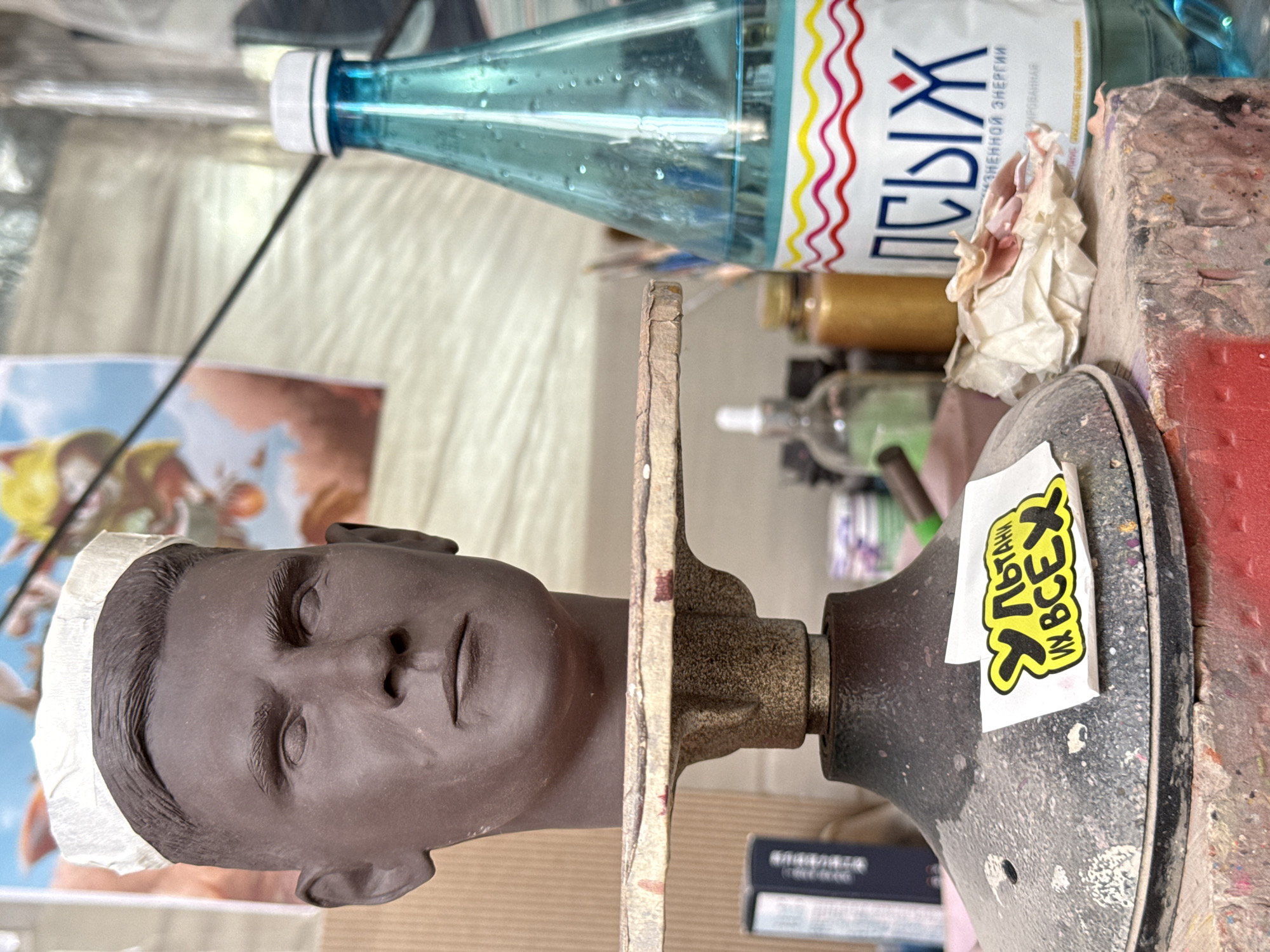 A tabletop scene features a clay sculpture of a person's head facing left, affixed to a turntable. A bottle of "Pishih" mineral water sits atop a cluttered surface. A sticker with Cyrillic text is adhered to the turntable's base. The location is likely Chaikovsky, Russia, based on the Cyrillic on the water bottle and sticker. The room seems to be a studio or workshop, given the presence of the sculpture and turntable.