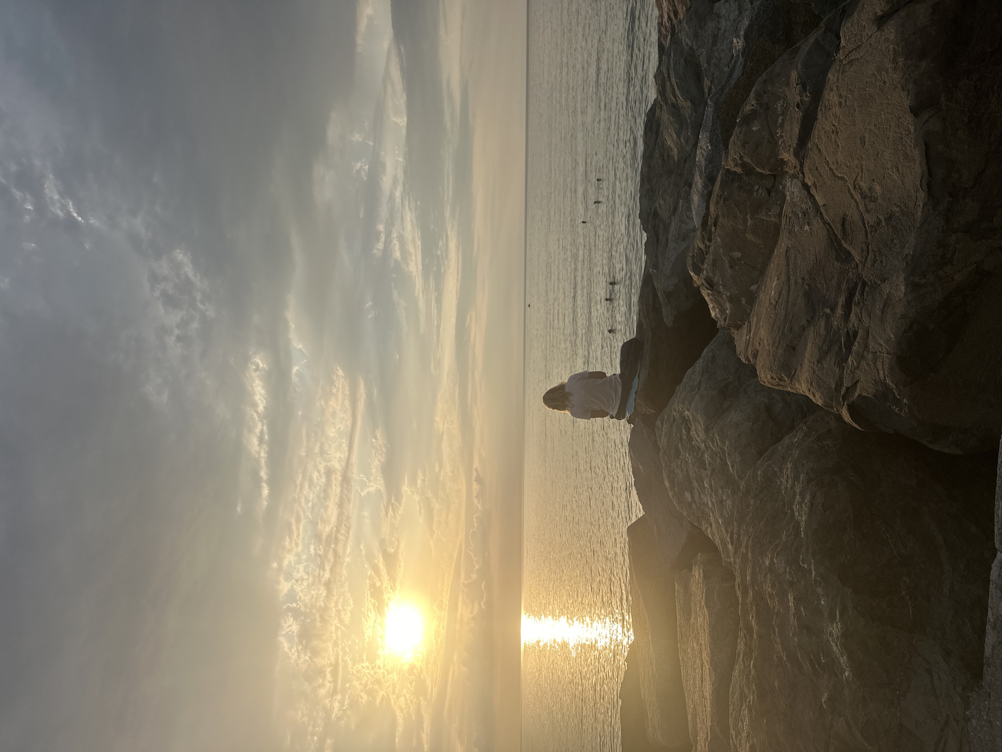 The image depicts a serene sunset scene at the Batumi Boulevard, a prominent public park and promenade in Batumi, Georgia. A lone individual, seen from behind, sits on large, dark rocks on the coastline. The person has shoulder-length, light-colored hair and wears a light-colored t-shirt and dark bottoms. They are seated on a blue mat or towel. The sun, a bright, luminous orb, is low in the sky, casting a golden, shimmering reflection on the calm sea. The sky is a mixture of soft grey and pale yellow clouds, creating a dramatic and atmospheric backdrop. In the distance, several other individuals are visible in the water, appearing to be swimming or enjoying the sea. A small boat is also visible on the horizon. The foreground is dominated by the textured, rough surfaces of the large rocks that form the breakwater. The overall impression is one of tranquility and natural beauty.