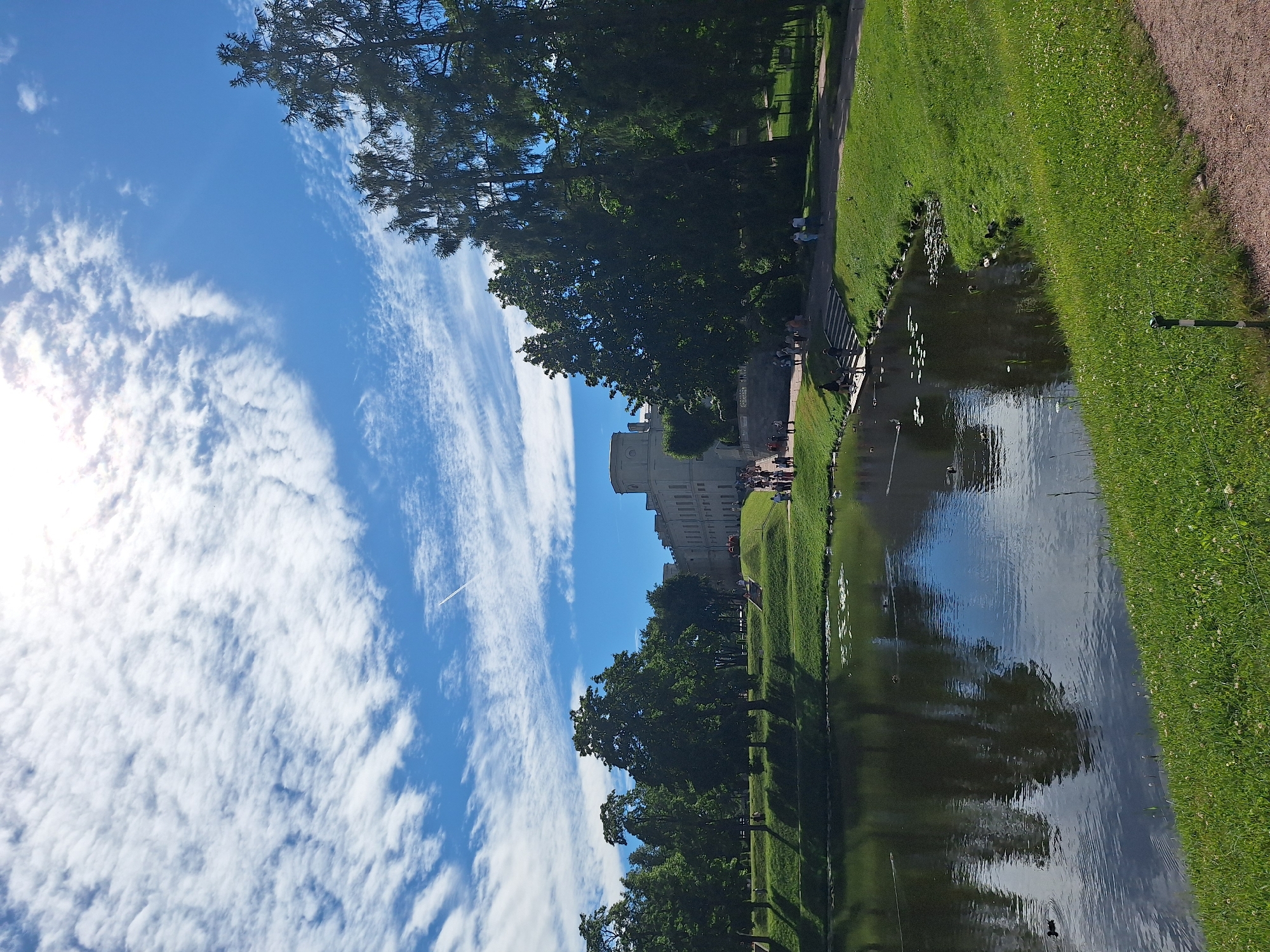 The image depicts a wide shot of the Gatchina Palace and its surrounding park in Gatchina, Russia. In the foreground, a calm body of water with lily pads and a few ducks is visible, reflecting the sky and trees. The water's edge is lined with lush green grass. Beyond the water, a gently sloping lawn leads up to a stately, light-colored palace structure with multiple wings and a distinctive round tower. Several people are seen walking along pathways and stairs leading to the palace. Tall, mature trees, primarily evergreens, flank the right side of the scene, casting shadows on the well-maintained grounds. The sky is a vibrant blue, adorned with scattered white clouds, and a faint contrail from an aircraft is visible. The overall scene conveys a peaceful, picturesque summer day in a historic park setting.