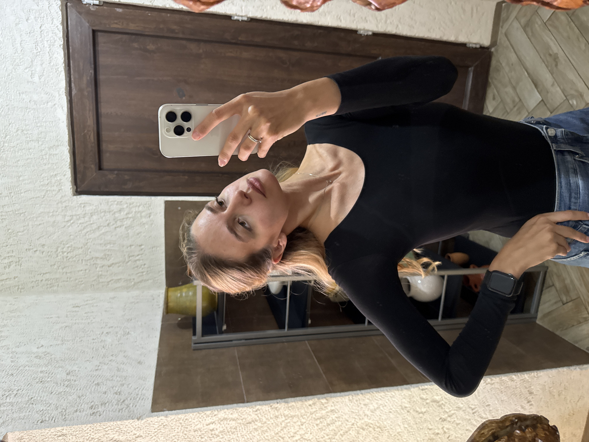 A young woman is captured in a mirror selfie, facing slightly away from the camera and looking into the reflection. Her head is tilted, and her expression is neutral to slightly smiling. She has light-colored hair, visible around her face and shoulders.
She is wearing a fitted black long-sleeved top or bodysuit and blue denim jeans. On her right hand, a simple ring is visible, and around her neck, she wears a delicate silver necklace with a small pendant. A black smartwatch is on her left wrist. She is holding a light-colored smartphone, likely an iPhone given its distinct triple camera setup, in her right hand, angling it towards the mirror to take the photo.
The background, visible through the mirror's reflection, consists of various elements. To her upper left, a dark wooden panel or door is present against a textured, light-colored wall. Further in the background, a shelving unit or cabinet can be seen holding decorative items, including a yellow vase-like object and a white ceramic piece. A light-colored patterned rug or carpet is visible at the bottom of the reflected scene.
A small, dark tattoo with stylized text or symbols, possibly "XUVY" or similar, is visible on the inner side of her right forearm, just above the wrist. The overall setting appears to be indoors, likely a home, given the combination of a mirror, a door, and decorative elements. The lighting suggests an indoor artificial light source or soft natural light.