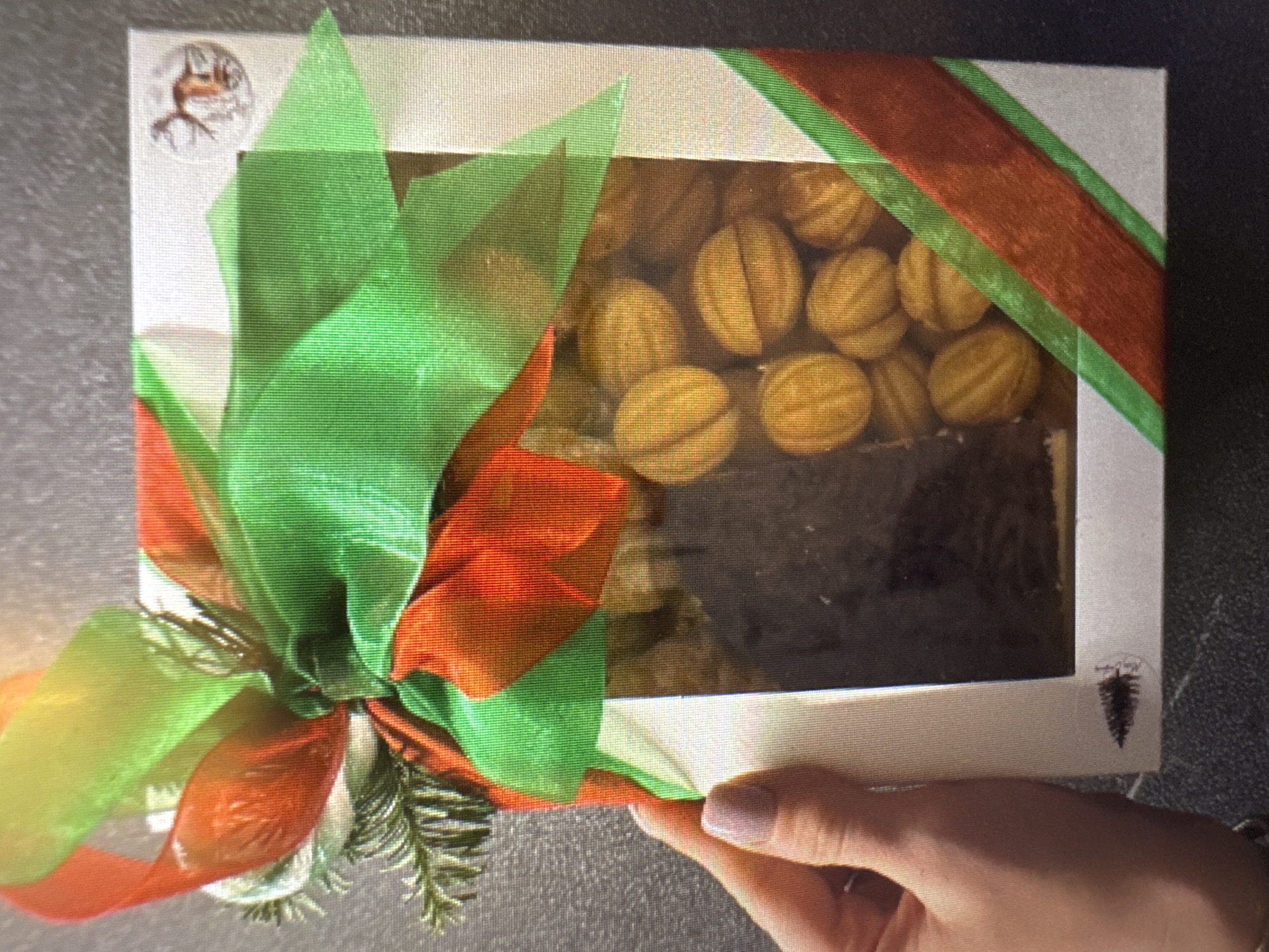 The image shows a rectangular gift box filled with small, round, golden-brown cookies. The box is decorated with a large bow made of red and green ribbons, along with a sprig of greenery. The box has a transparent window revealing the cookies inside, which appear to be a type of walnut-shaped shortbread or biscuit, possibly "Gozna Laski" from Eastern Europe. The box also has diagonal red and green ribbon details on the sides. A person's hand, with a light pink fingernail polish, is visible on the left side of the image, holding the box. The background appears to be a dark, textured surface, possibly a countertop or table. There is no information in the image to suggest its location in the city of Murcia, Spain.