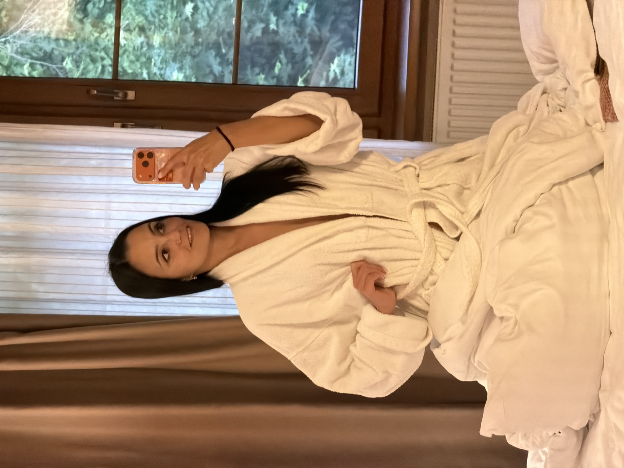 A woman with long, dark hair parted in the middle is seated on a bed, holding a smartphone in her left hand to take a selfie. She is wearing a white terrycloth bathrobe, loosely tied at the waist, revealing a slight opening at the chest. Her left arm is extended, holding an orange-cased smartphone featuring a three-lens camera module. A dark elastic band is present on her left wrist. Her right hand lightly grasps a section of the bathrobe fabric near her hip. She exhibits a subtle smile and directs her gaze towards the phone. The bed is covered with white sheets and a white duvet that appears rumpled. To the viewer's left, a heavy brown curtain is partially drawn across a window, behind which sheer white curtains are visible. To the right, a dark brown wooden-framed window is visible, through which dense green foliage can be seen. A white, ribbed radiator is situated below the window frame on the right side of the bed. The scene is located in an indoor room in Svetlogorsk, Russia.