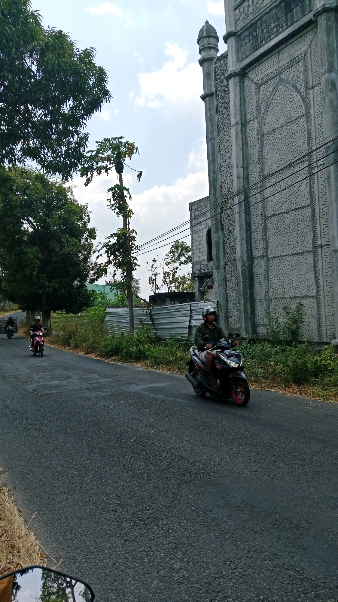 This is a slightly low-angle, eye-level shot of a street in Sumberagung, Indonesia. The scene is dominated by a large, ornate concrete building on the right, possibly a mosque, featuring intricate geometric carvings. A paved road runs diagonally across the frame from the lower left to the middle right, with a person riding a black scooter on it, heading towards the viewer. This individual wears a helmet and a dark green jacket. To the left of the road, lush green trees and vegetation are visible, with a tall, slender tree bearing fruit in the mid-ground. Further down the road, two more scooters are seen, one red and one black. The sky is partly cloudy with patches of blue.