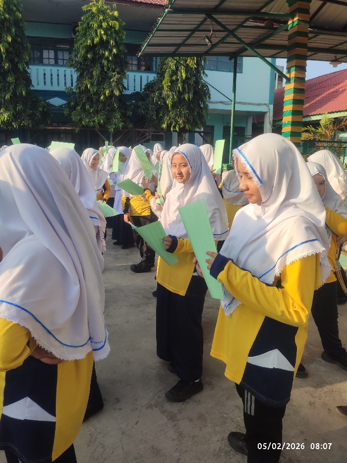 A group of young women wearing white hijabs and yellow and black school uniforms stand outside holding green paper. They are lined up in rows and some are looking at their papers while others look towards the camera. In the background are trees and a building with a green and white color scheme. The photo appears to have been taken in the morning as indicated by the date and time stamp on the bottom right.