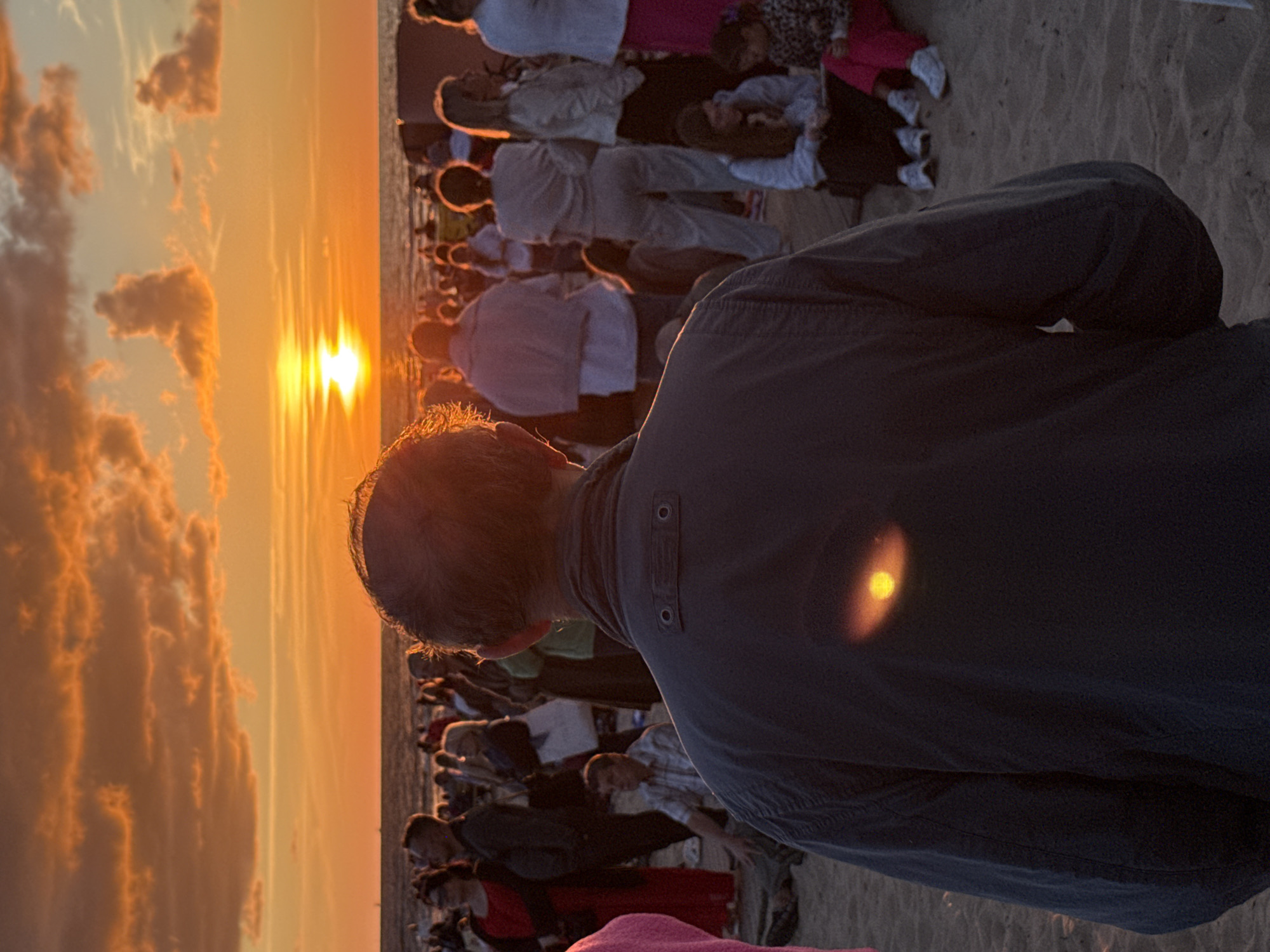 The image depicts a crowd of people on a sandy beach at sunset. The sky is filled with vibrant orange and yellow clouds, with the sun prominently visible near the horizon, casting a warm glow over the scene. Several individuals are visible in the foreground and background, some facing the sunset, others interacting with each other. The main subject in the foreground is a person seen from behind, wearing a dark jacket. In the middle ground, there are more people standing and sitting, some dressed in casual attire. The environment suggests a public gathering or event taking place outdoors. There is no information in the image to determine the specific location in the city of Riga, Latvia.