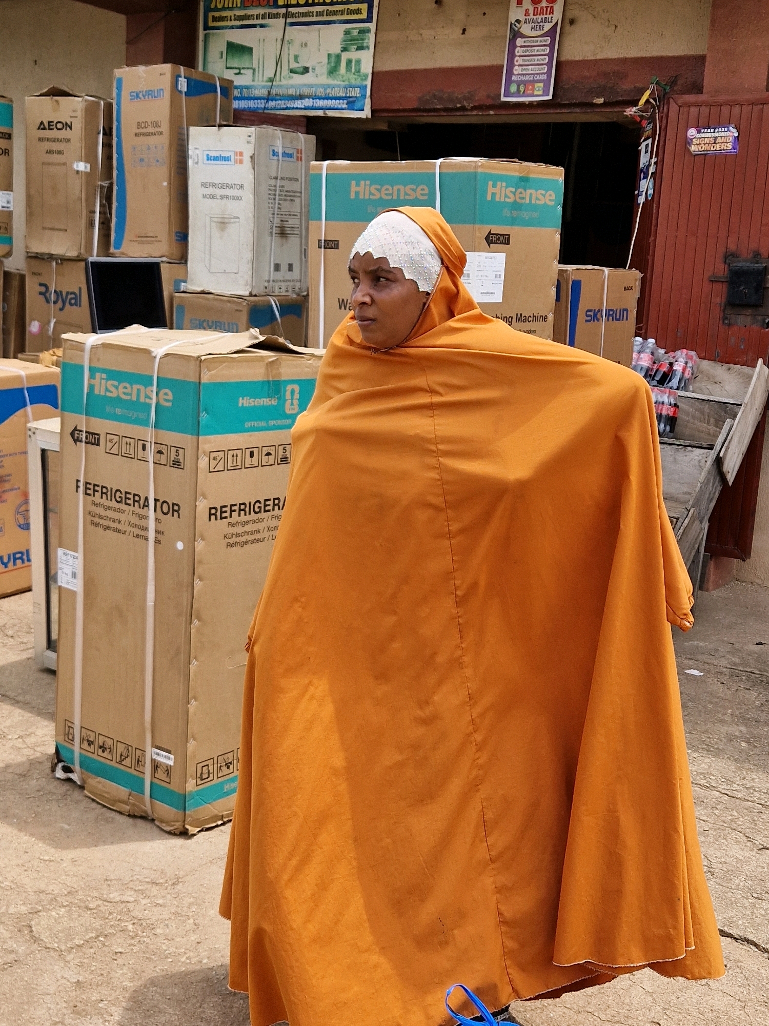 The image depicts an outdoor market scene in Jos, Nigeria, with a prominent display of boxed appliances, primarily refrigerators. In the foreground, a woman wearing a white headscarf and an orange garment that drapes over her body is the central subject. She is looking to her right, her face partially obscured by her attire. Behind her, stacked cardboard boxes are clearly labeled with brand names such as "Hisense," "Skyrun," "Scanfrost," and "Royal." Some of these boxes are open, revealing the contents or packaging. The boxes suggest a retail or wholesale environment for home appliances. In the background, a building with a reddish-brown facade and a shop entrance is visible, with signs and posters displayed on the wall. One sign advertises "Year 2020 Commissioned Signs and Wonders." To the right, a wooden structure holds several bottles of what appear to be soft drinks. The ground is a rough, paved surface, consistent with a market area. The overall impression is one of commerce, with a focus on the distribution of goods in a Nigerian urban setting.