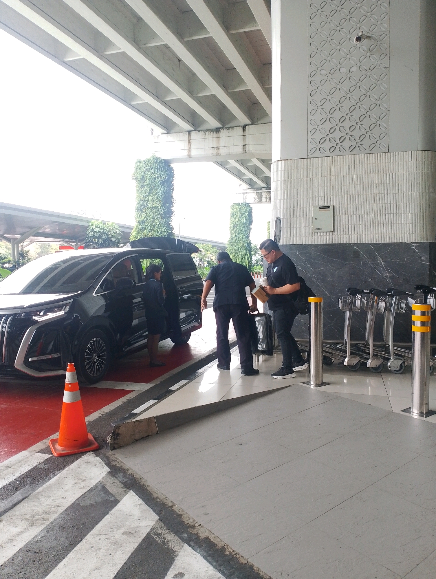 An outdoor scene in Tangerang, Indonesia, shows two men in black uniforms and one woman in a dark blue uniform near a black luxury van with its rear door open. The van is parked on a red surface with white lines, adjacent to a grey tiled walkway. An orange traffic cone is positioned on the ground near the van. In the background, a white building with decorative patterns and a security camera is visible, along with several luggage trolleys. Above the scene, a concrete overpass with exposed beams is present. The lighting suggests it is daytime.