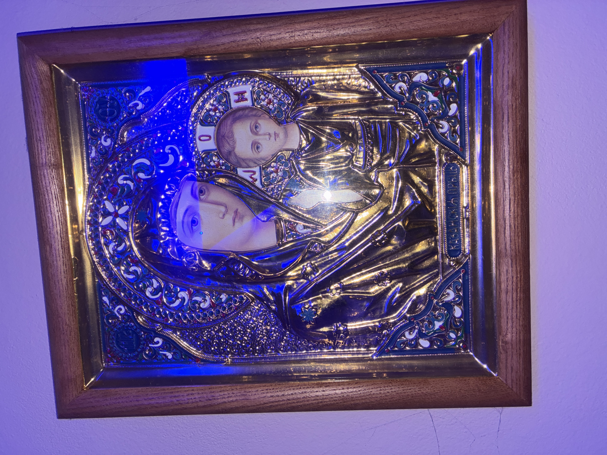 An ornate, framed icon of the Virgin Mary and child Jesus hangs on a plain white wall. The icon is rendered in a metallic, likely gold, material with intricate detailing. The Virgin Mary is depicted with a blue jewel on her forehead and wears a golden veil. The child Jesus is also depicted in golden robes. Both figures have halos surrounding their heads, with Greek letters inscribed within them. The frame of the icon is wooden and appears to be natural in color. The wall exhibits some subtle texture and faint lines. Blue lighting illuminates the scene from the upper left, casting a purplish hue on the wall and a blue reflection on the icon. The overall impression is one of religious art displayed in a domestic setting. The location is not discernible from the image.