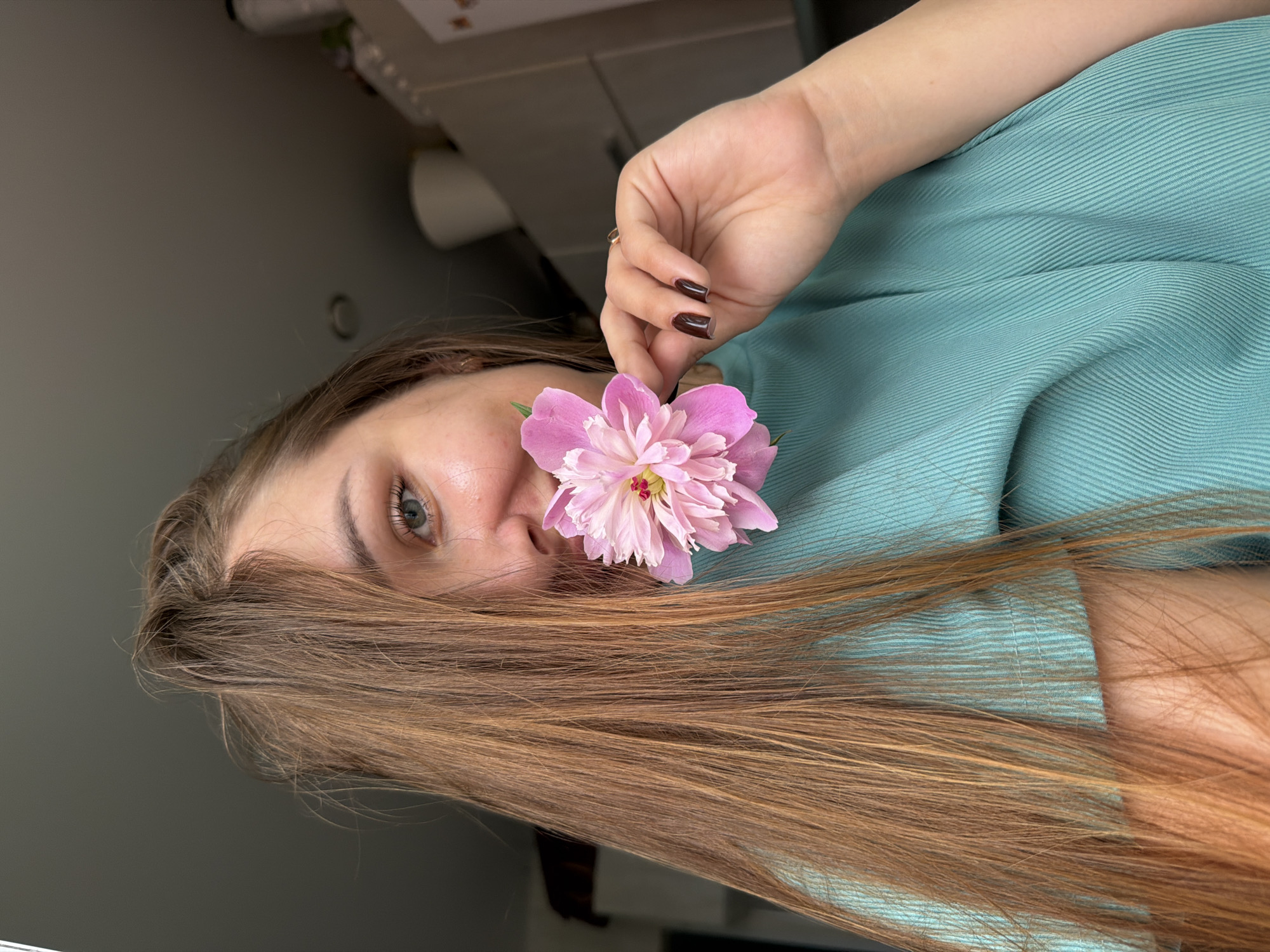 A young woman with long, brown hair and green eyes is holding a pink peony flower up to her face, partially obscuring her mouth. She is wearing a teal-colored, ribbed t-shirt. Her left hand, with dark nail polish, is holding the stem of the flower, and a ring is visible on her right ring finger. She appears to be indoors, with a gray ceiling and cabinets visible in the background. The lighting suggests it is daytime. The image does not provide any specific information about the location in the city of Chaikovsky, Russia.