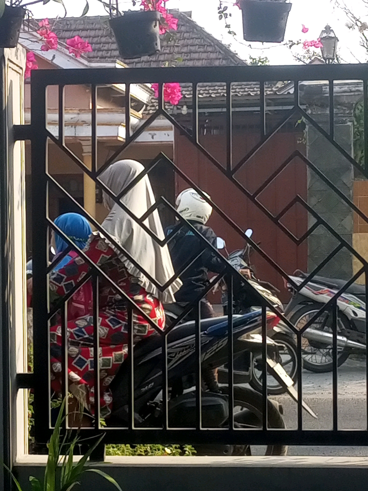 A scene captured through a black metal gate in Ngasem, Indonesia, shows two people on a motorcycle. The person in the front, wearing a white headscarf and a red and white patterned dress, is likely a passenger. Behind them, the driver, wearing a white helmet and a dark jacket, is seated on the motorcycle. The motorcycle is positioned on a paved surface, with another motorcycle visible to the right. The background includes a building with a red-brown wall and a tiled roof, adorned with hanging plants bearing pink flowers. A house with a white facade and windows is also visible. The image is taken during daylight.