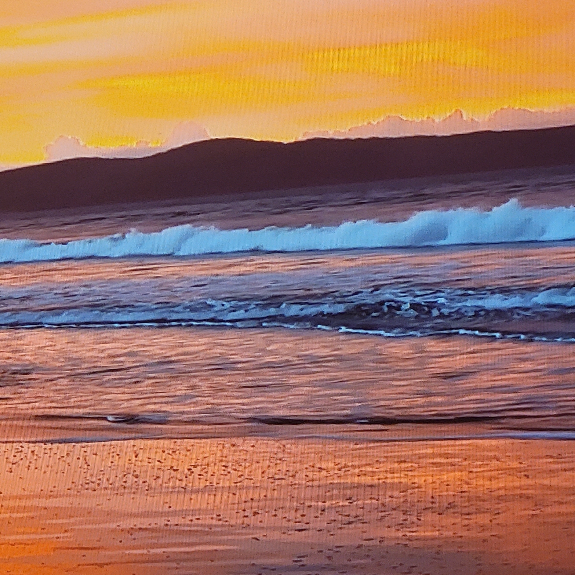 The image depicts a beach scene at sunset. The sky is a gradient of orange and yellow. A dark, undulating landmass is silhouetted against the horizon. Waves are breaking on the shore, with white foam cresting above the blue water. The wet sand reflects the colors of the sky.
The scene is a generic beach landscape and cannot be accurately pinpointed in Yerevan, Armenia, as Yerevan is a landlocked city and does not have a coastline.
