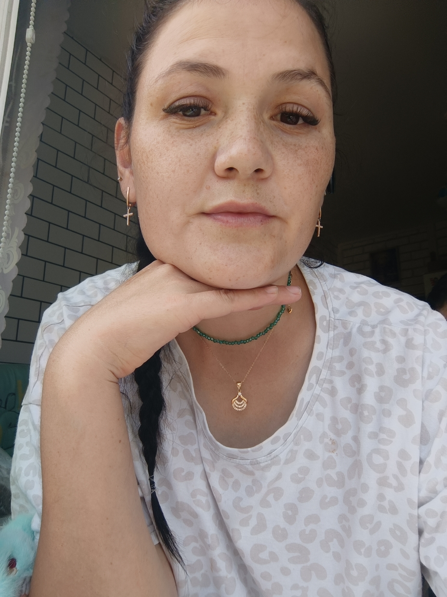 A medium shot captures a woman with dark hair pulled back and a braid extending down her left side. She rests her chin on her right hand, her gaze directed slightly off-camera. She is wearing a light-colored shirt with a subtle leopard print and two necklaces: a beaded green one and a delicate gold pendant. Small gold cross earrings adorn her ears. The background features a white brick-patterned wall to the left and a dark, indistinct area to the right, suggesting an indoor setting. There is no discernible location in the city of Chaikovsky, Russia.