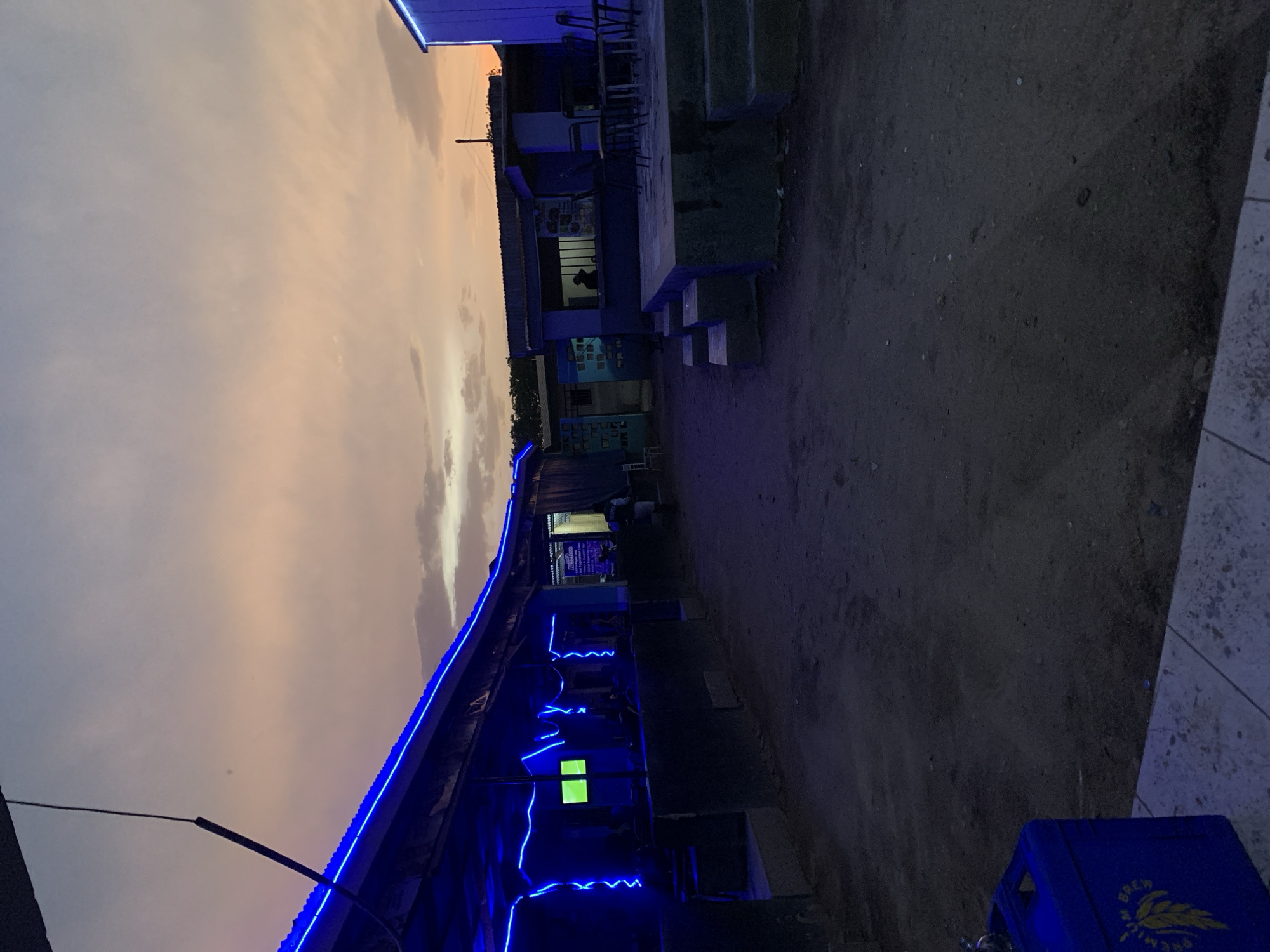 The image depicts a night scene in Kabong, Nigeria. The sky is a mix of grey and orange hues. A building is decorated with blue neon lights, including a glowing screen. People are visible inside the building. The ground is a dark, dirt-like surface. A blue crate is in the foreground. The camera appears to be oriented upwards.