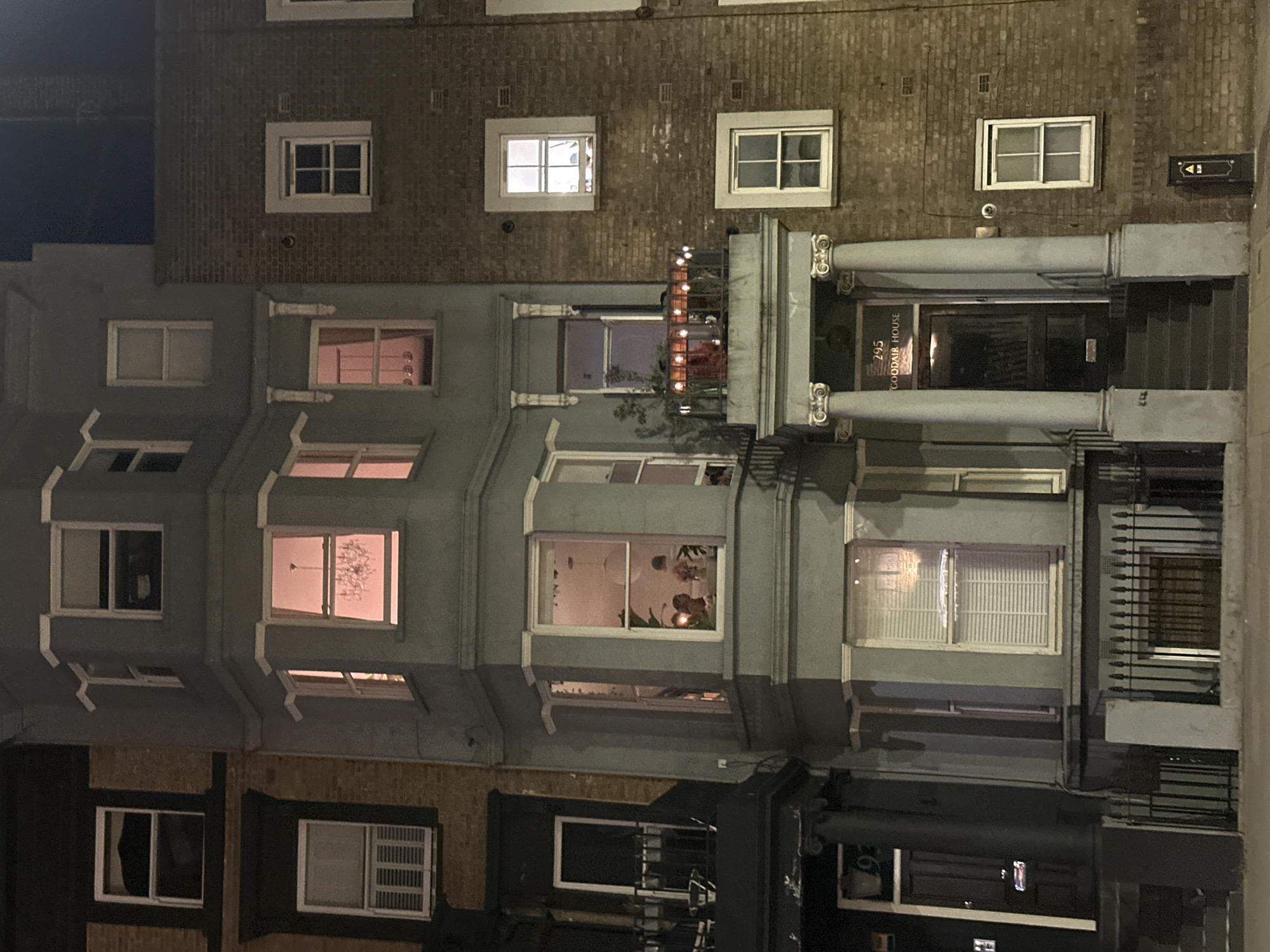 A nighttime photograph depicts a row of multi-story buildings. The buildings are primarily brick and grey. The front facade of one building has an entranceway framed by ornate columns. The address "295 GOODAIR HOUSE" is visible above the door. Various windows are lit up on the buildings. Some windows display interior objects such as a chandelier and plants. One building has a small balcony with string lights. The street level appears dark, with shopfronts and wrought iron bars visible. The location is likely London, United Kingdom, based on the architectural style.