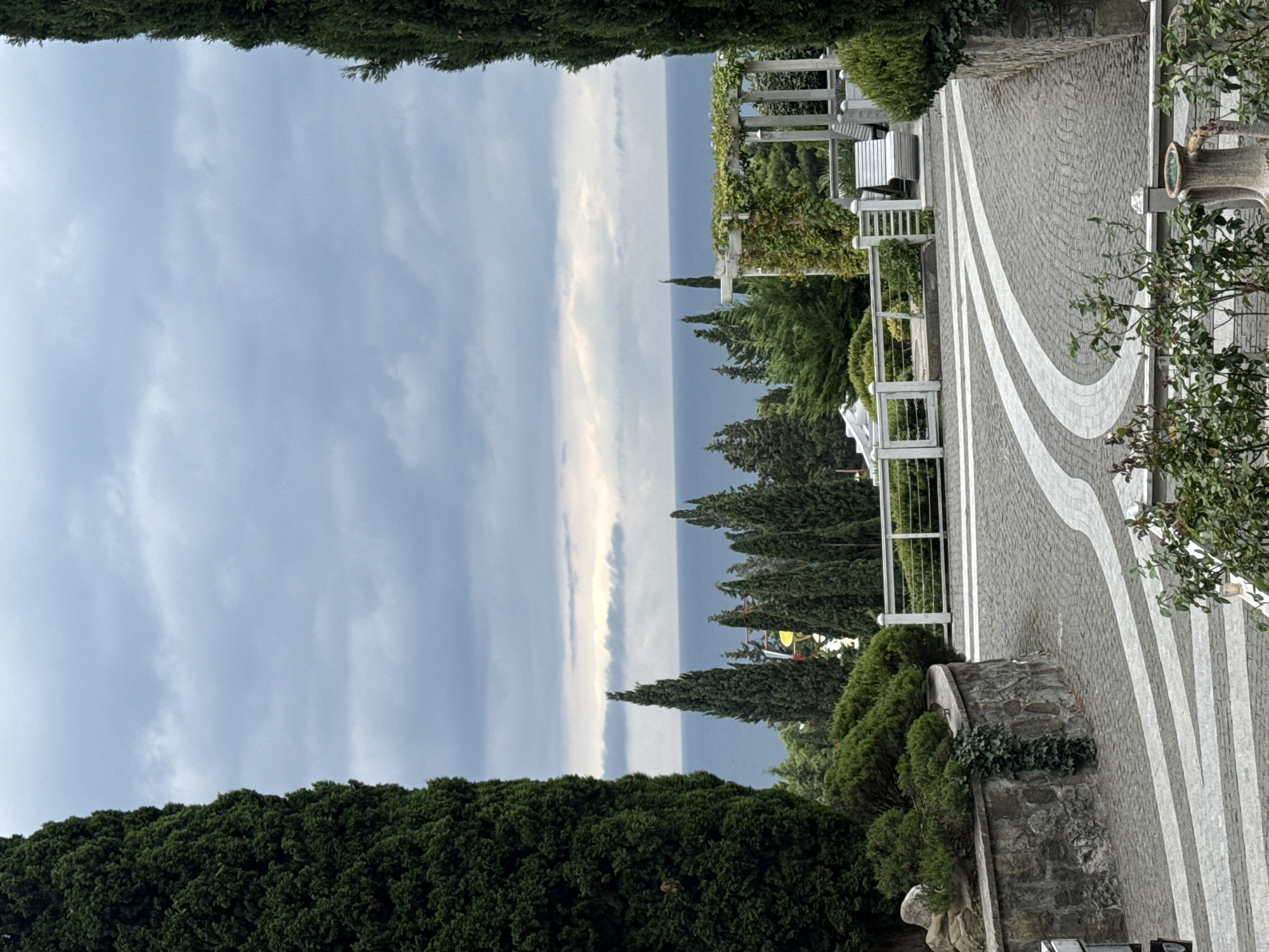 The image shows a view of the Black Sea from a landscaped area in Yalta, Ukraine. The foreground features a cobblestone pathway with white curved lines, bordered by stone walls and manicured greenery. Tall cypress trees frame the scene on either side, with a white fence and a wooden pergola with climbing plants visible in the middle ground. Beyond the greenery, the calm blue sea stretches to the horizon, under a cloudy sky. The overall impression is one of a well-maintained park or garden area overlooking the coastline.