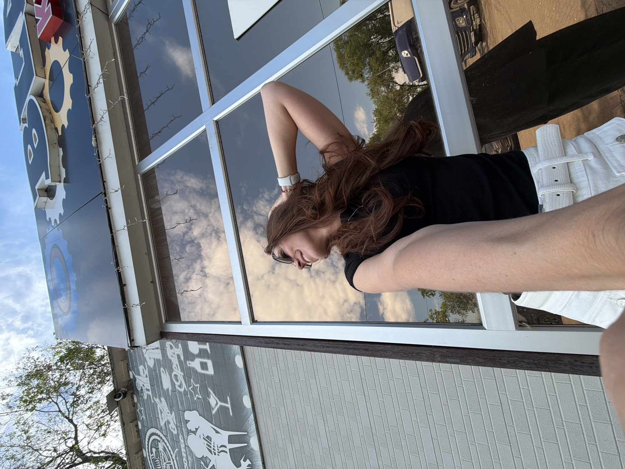 A woman with long, reddish-brown hair, wearing sunglasses and a black t-shirt, poses for a selfie in front of a building with large windows that reflect the sky and surrounding environment. She is holding her left hand behind her head and her right arm is extended, holding the camera. She is wearing a white belt and white shorts. The building has a sign above the windows, with the word "POLO" visible in large letters. The background shows a brick wall with decorative designs and a tree. In the reflection of the window, a blue truck and trees are visible. The scene appears to be in an outdoor urban setting.