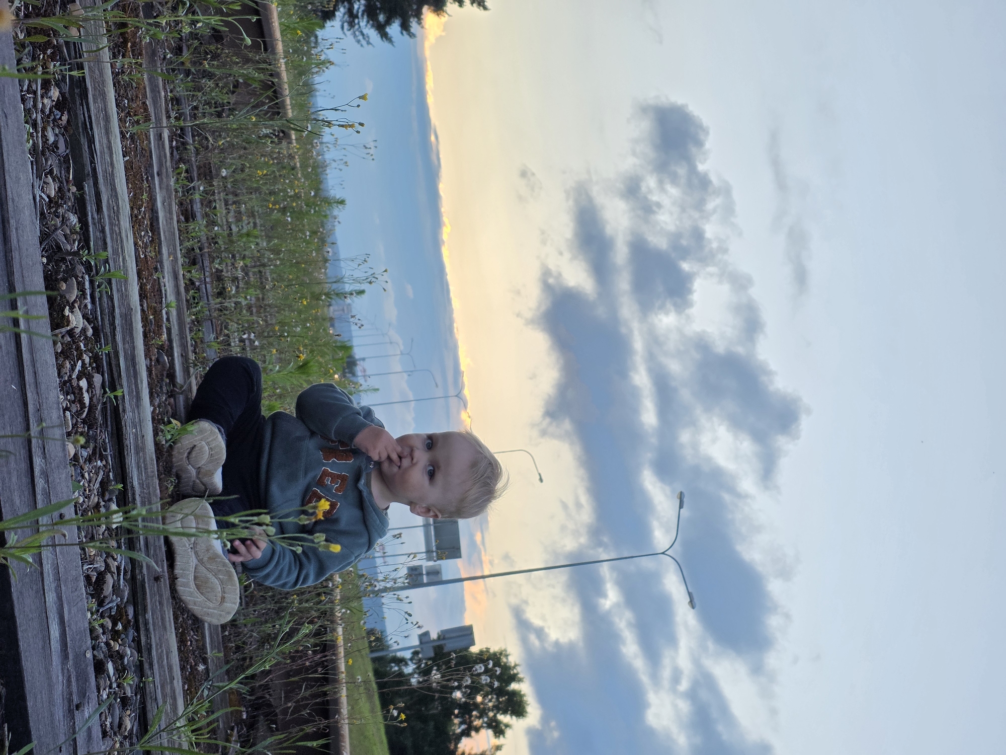 A young child with blonde hair sits on abandoned railway tracks amidst overgrown weeds and wildflowers, with a yellow flower near their face. The child is wearing a grey hooded sweatshirt and black pants, with one hand touching their mouth and the other resting on their leg. In the background, streetlights line a road, and the sky is filled with scattered clouds, with a band of orange and yellow light near the horizon, suggesting sunset. The scene appears to be in an urban or suburban area, possibly on the outskirts of Chaikovsky, Russia, given the presence of infrastructure like streetlights and railway tracks.
