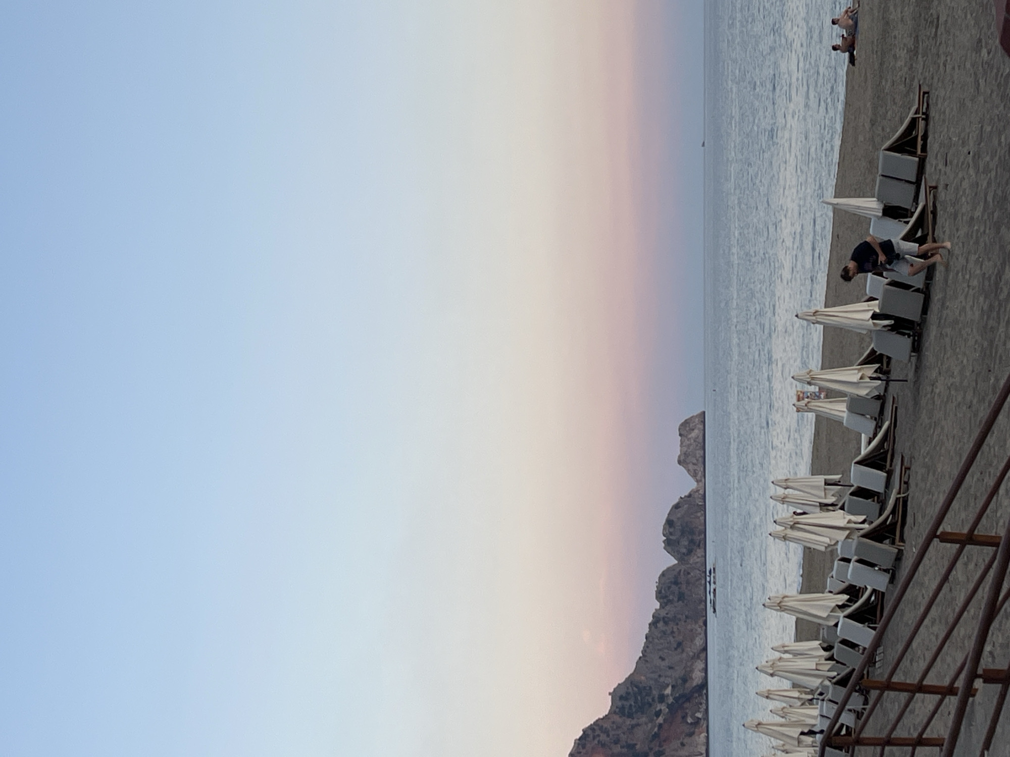 This image presents a serene beach scene captured during what appears to be late afternoon or early evening, indicated by the soft, gradient sky. The sky transitions from a light blue at the top left to gentle shades of pale pink and peach closer to the horizon, suggesting either sunrise or sunset. The photograph depicts a long stretch of sandy and pebbly beach meeting a calm, placid sea.
Along the beach, numerous white beach chairs with folded white umbrellas are neatly lined up. In the foreground, one man, dressed in a dark t-shirt and light shorts, stands near an umbrella, seemingly adjusting something or observing the surroundings. Further down the beach, closer to the water's edge, two or three individuals are seen relaxing, lying directly on the sand. Another person is seated on a sunbed further in the distance.
In the mid-ground, rugged, rocky mountains or cliffs rise from the sea, framing the left side of the scene and adding a dramatic natural element to the landscape. A small boat is visible on the water, positioned near these distant landforms. On the immediate right side of the foreground, a dark metal railing suggests the image was taken from an elevated viewpoint or a structure overlooking the beach. The water appears calm, gently reflecting the subdued light of the sky. The overall mood conveyed is peaceful and relaxing, typical of a resort beach at dusk. No visible text is present in the image.