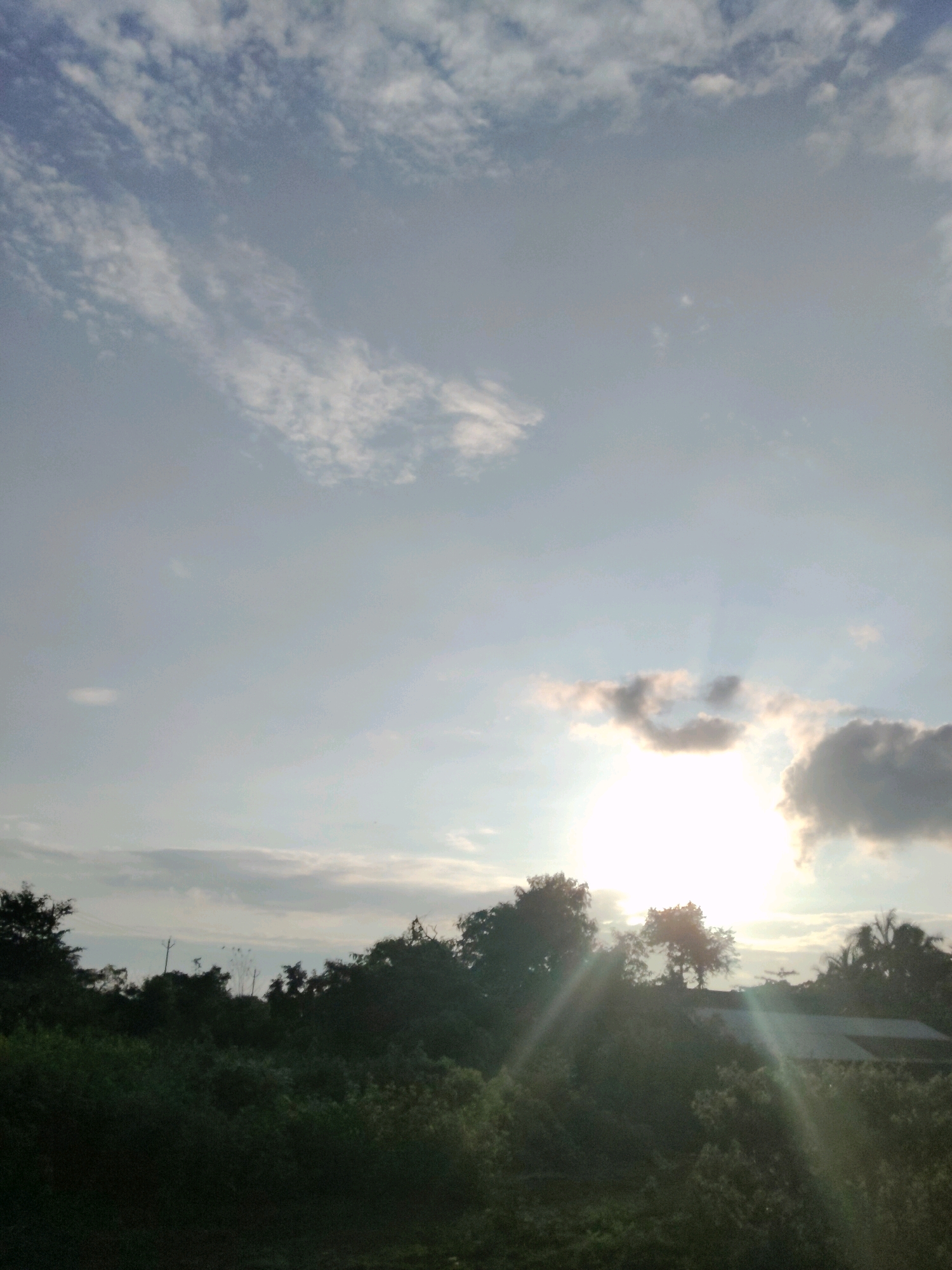 A bright sun shines through scattered clouds, casting rays over a dense, dark green landscape of trees and bushes. The sun is partially obscured by a cloud formation that resembles a smiling face. In the background, a roof of a building is visible through the foliage, suggesting a rural or semi-rural setting. The sky is a pale blue with wispy white clouds. The scene appears to be taken from an elevated position looking out over the landscape. The lighting suggests either sunrise or sunset. There are no people or animals visible in the image. The location is described as being in Vyara, India, which is a city in the Tapi district of Gujarat.